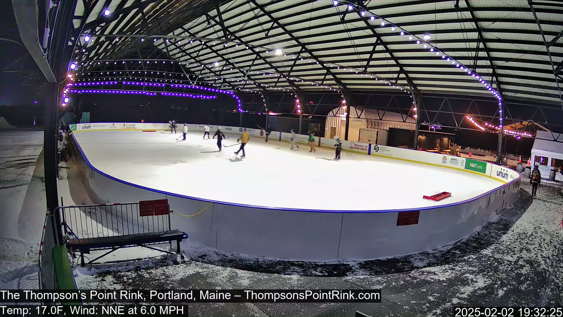 An outdoor ice rink at night, under a metal structure with string lights, is partially covered in snow with several people ice skating.