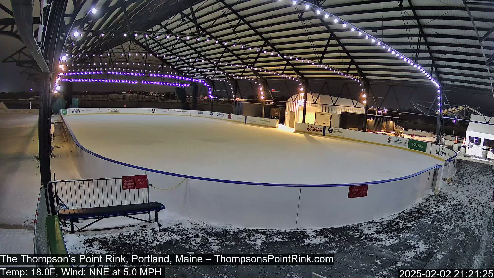 An outdoor ice rink, illuminated at night under a metal structure with string lights, is partially covered in snow.