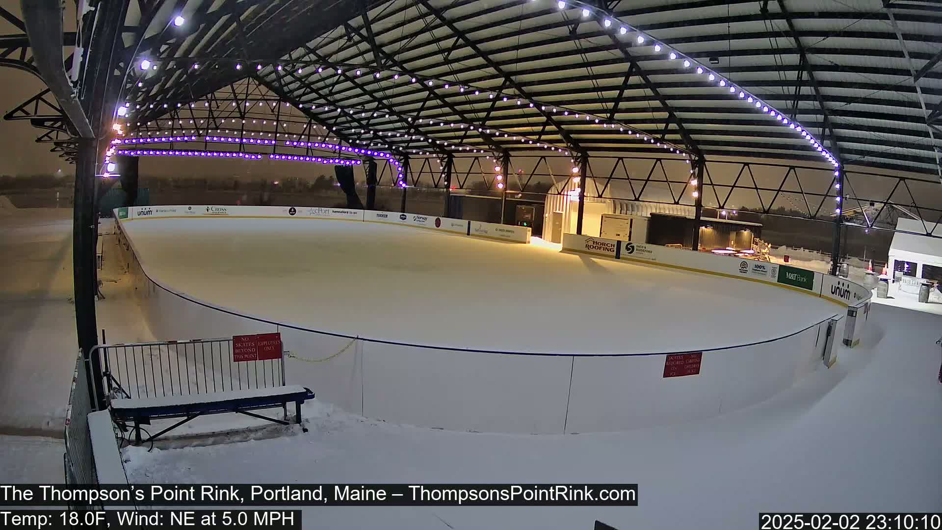 An outdoor ice rink, covered by a metal structure with string lights, is lightly snowing at night.