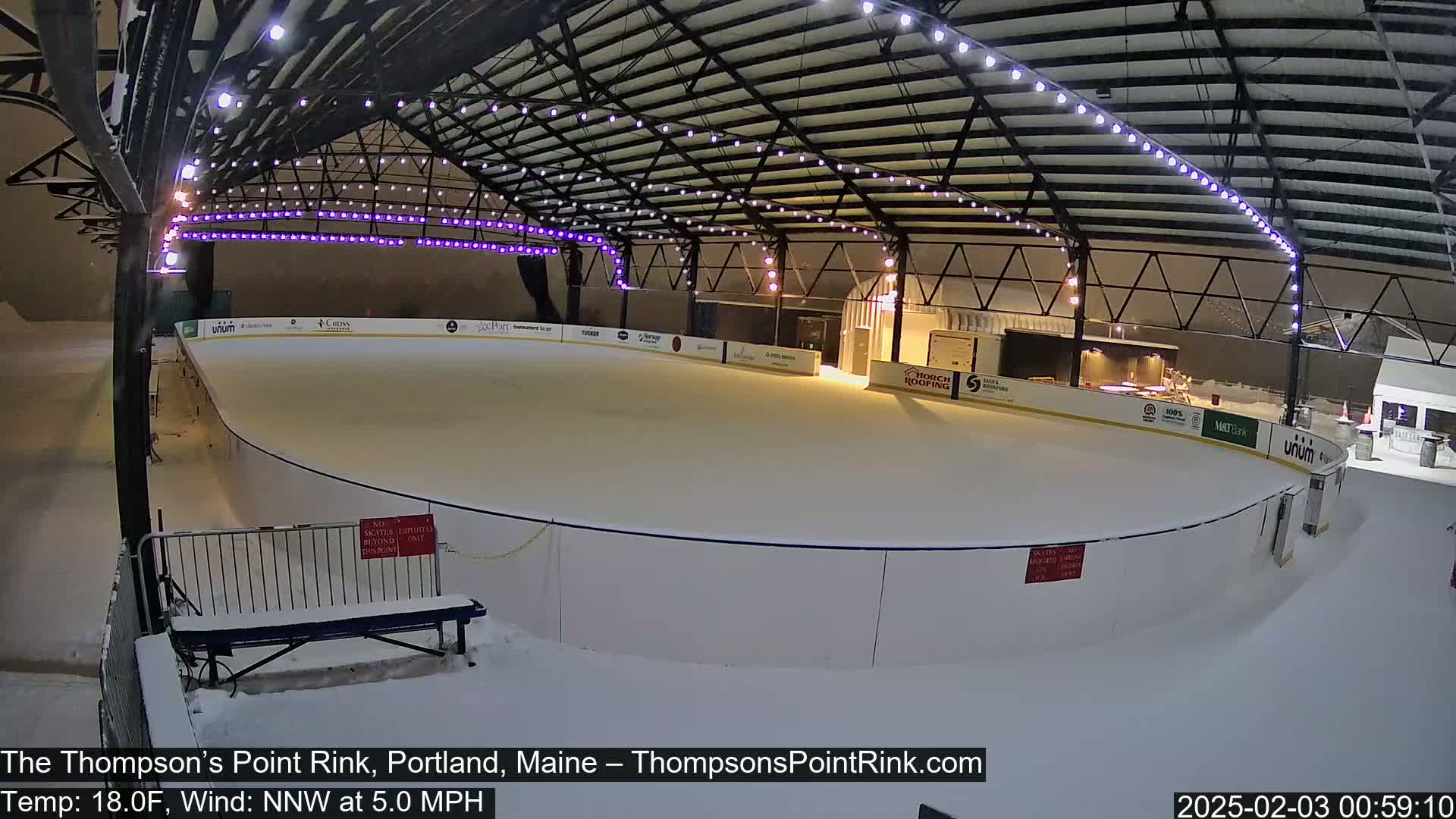 A snow-covered outdoor ice rink is illuminated under a metal structure with string lights, during a nighttime snowfall with a temperature of 18°F and a north-northwest wind of 5 mph.