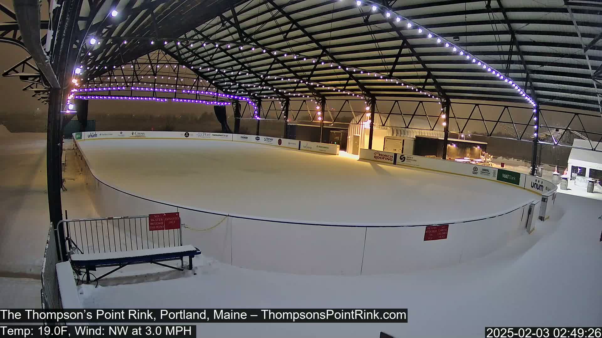 A snow-covered outdoor ice rink is illuminated at night under a metal structure with string lights.