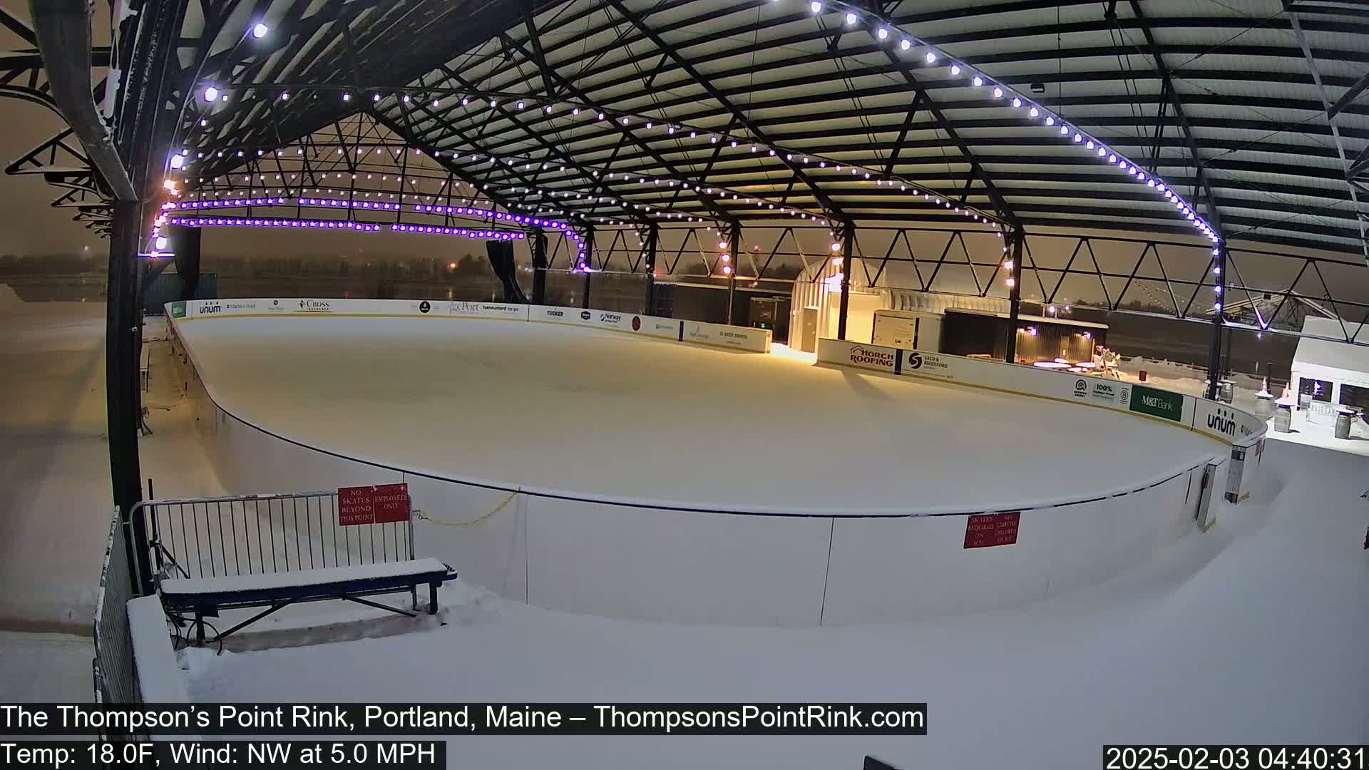 A snow-covered outdoor ice rink sits under a metal structure with string lights, during a nighttime snowfall with a northwest wind.