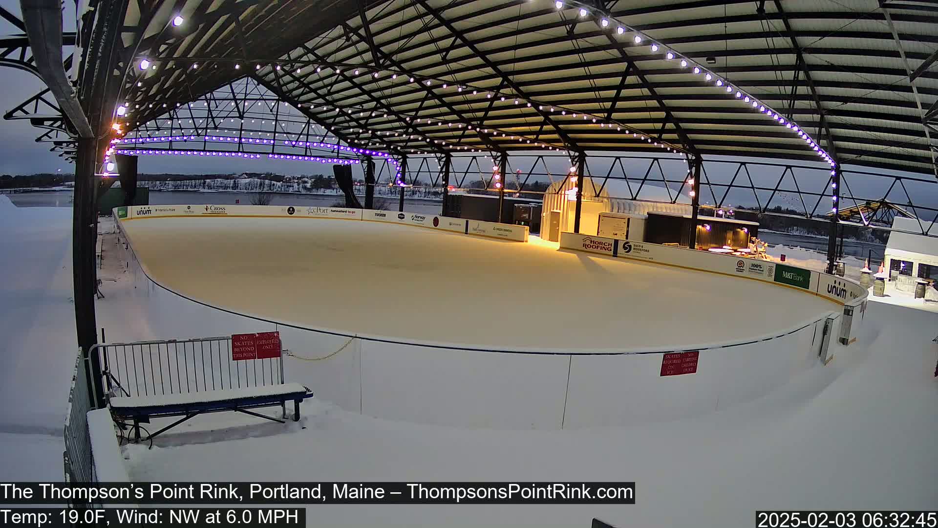 An outdoor ice rink under a metal structure with lights, surrounded by snow in twilight conditions.