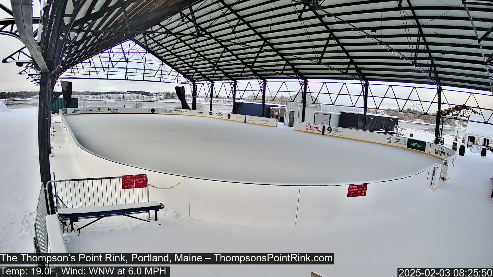 A snow-covered outdoor ice rink sits under a metal-framed structure.