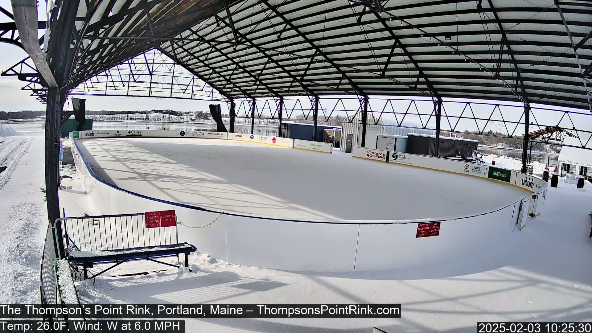 An outdoor ice rink, covered by a metal structure, is covered in snow on a sunny, windy day.