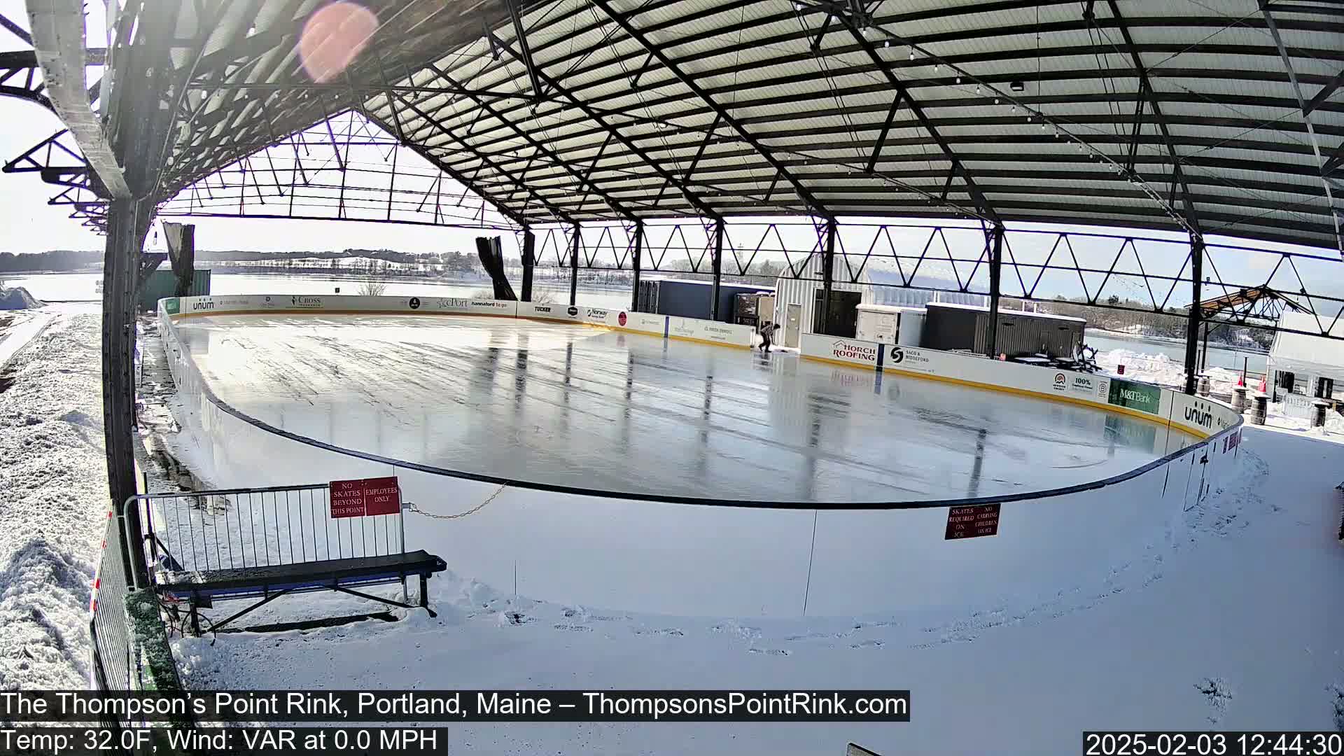 A sunny outdoor ice rink, covered by a metal structure, is partially surrounded by snow.