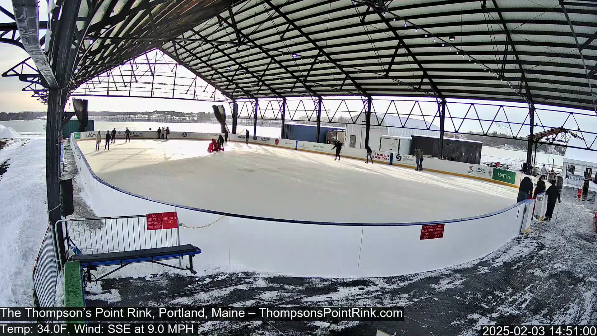 An outdoor ice rink, partially covered by a metal structure, is surrounded by snow, with several people ice skating on a sunny day.