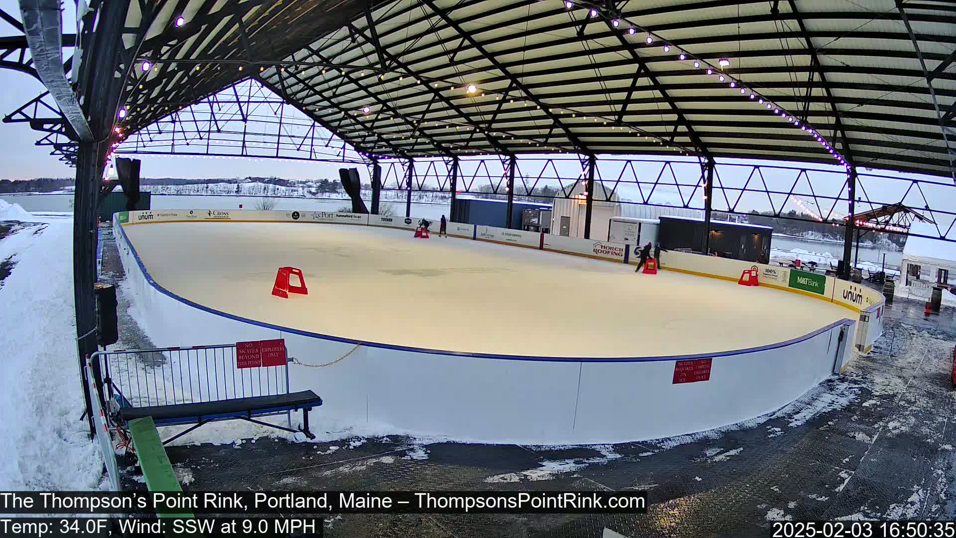 A partially covered outdoor ice rink sits mostly empty under an overcast sky, with a light dusting of snow on the ground and surrounding area.