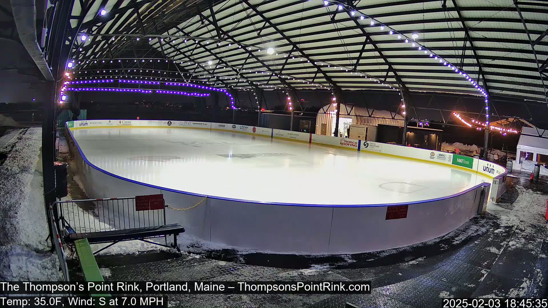An outdoor ice rink, illuminated at night, is surrounded by snow with a light south wind.