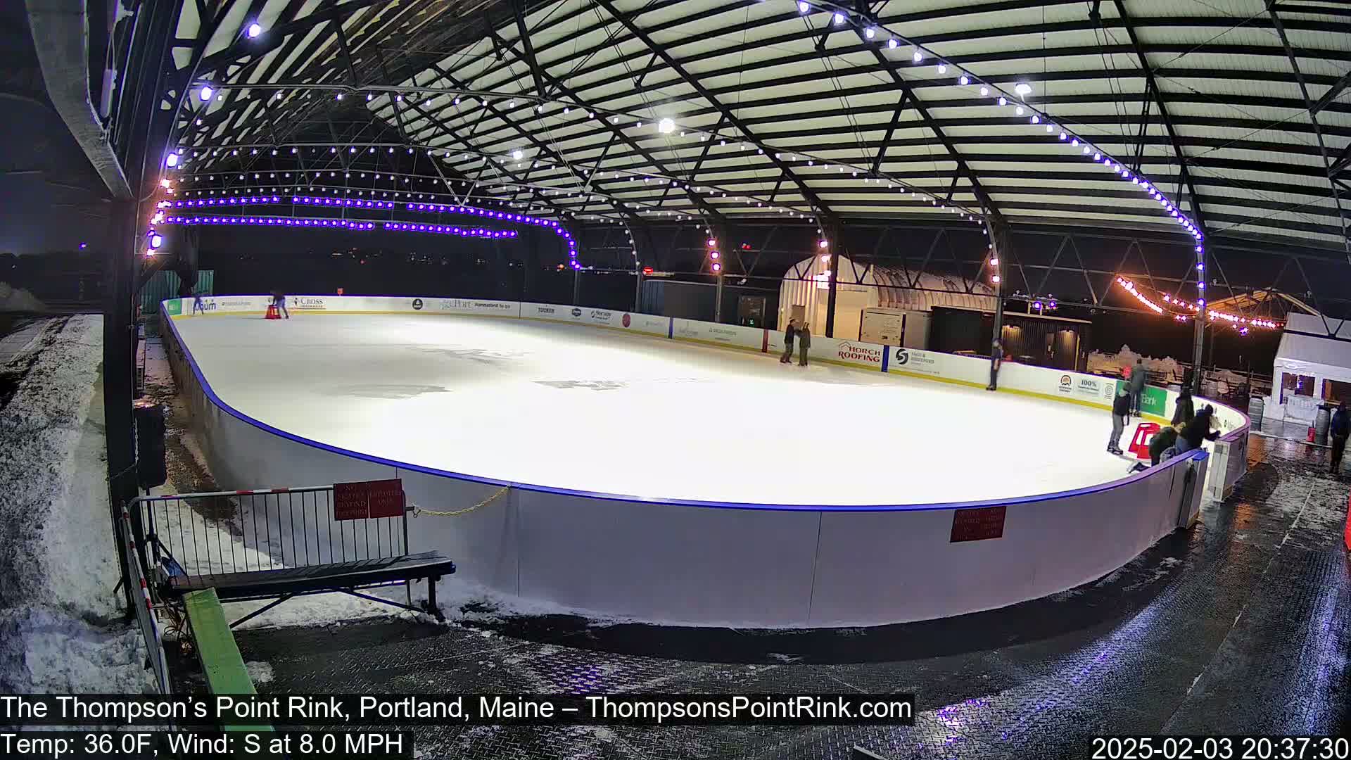 An outdoor ice rink, illuminated at night with string lights, under a metal roof, with some people ice skating and others standing around; it is 36°F with a south wind at 8 mph.