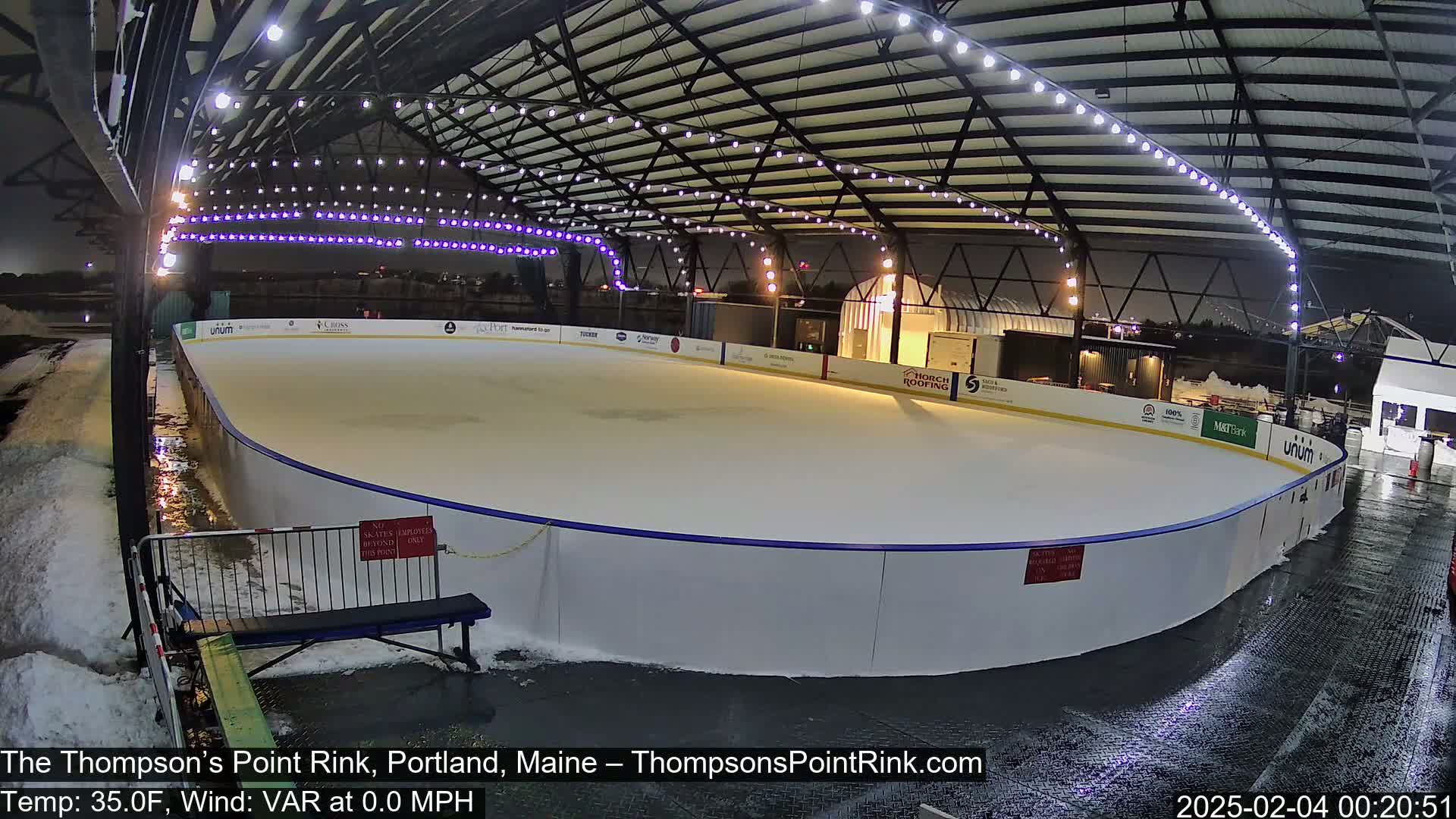 An outdoor ice rink, lit at night under a metal structure, is surrounded by snow and wet pavement.