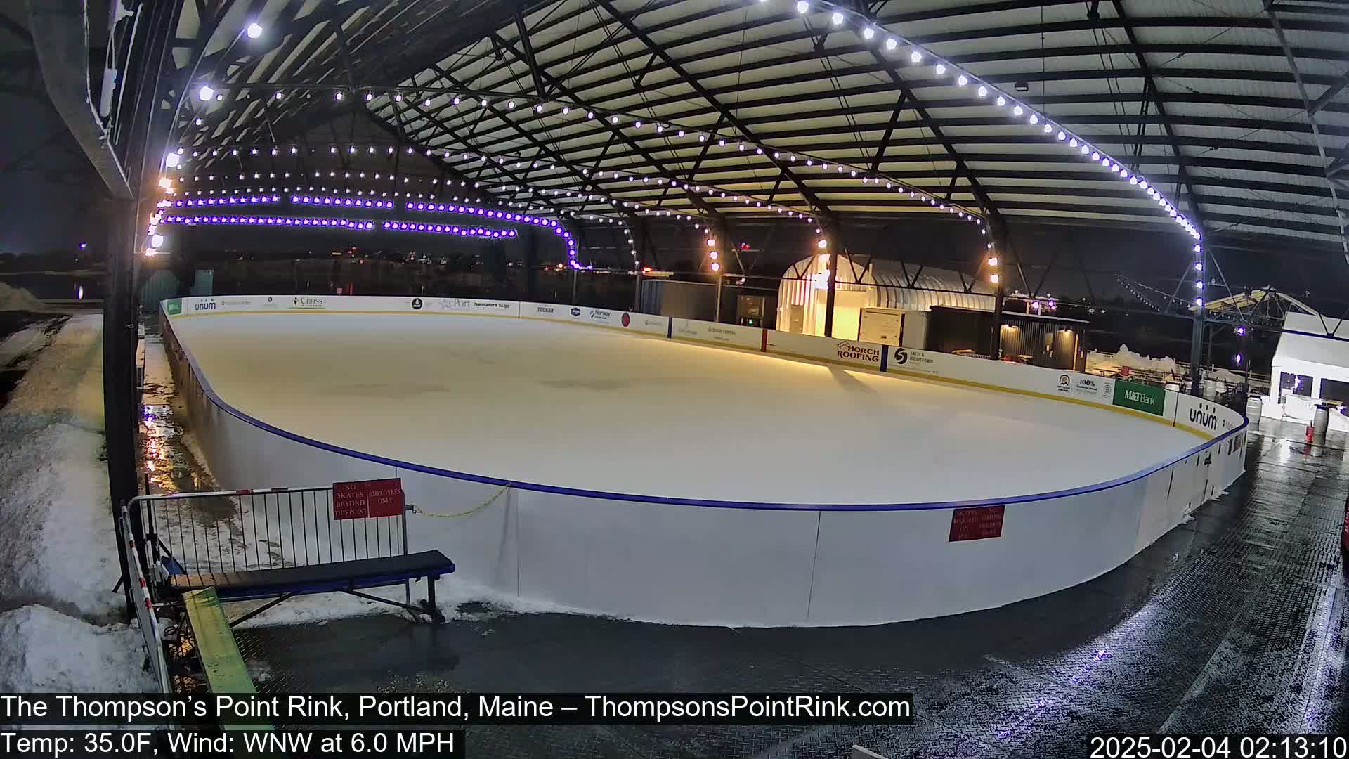 An outdoor ice rink is lit at night under a metal structure, with wet ground surrounding it and snow visible in the background.