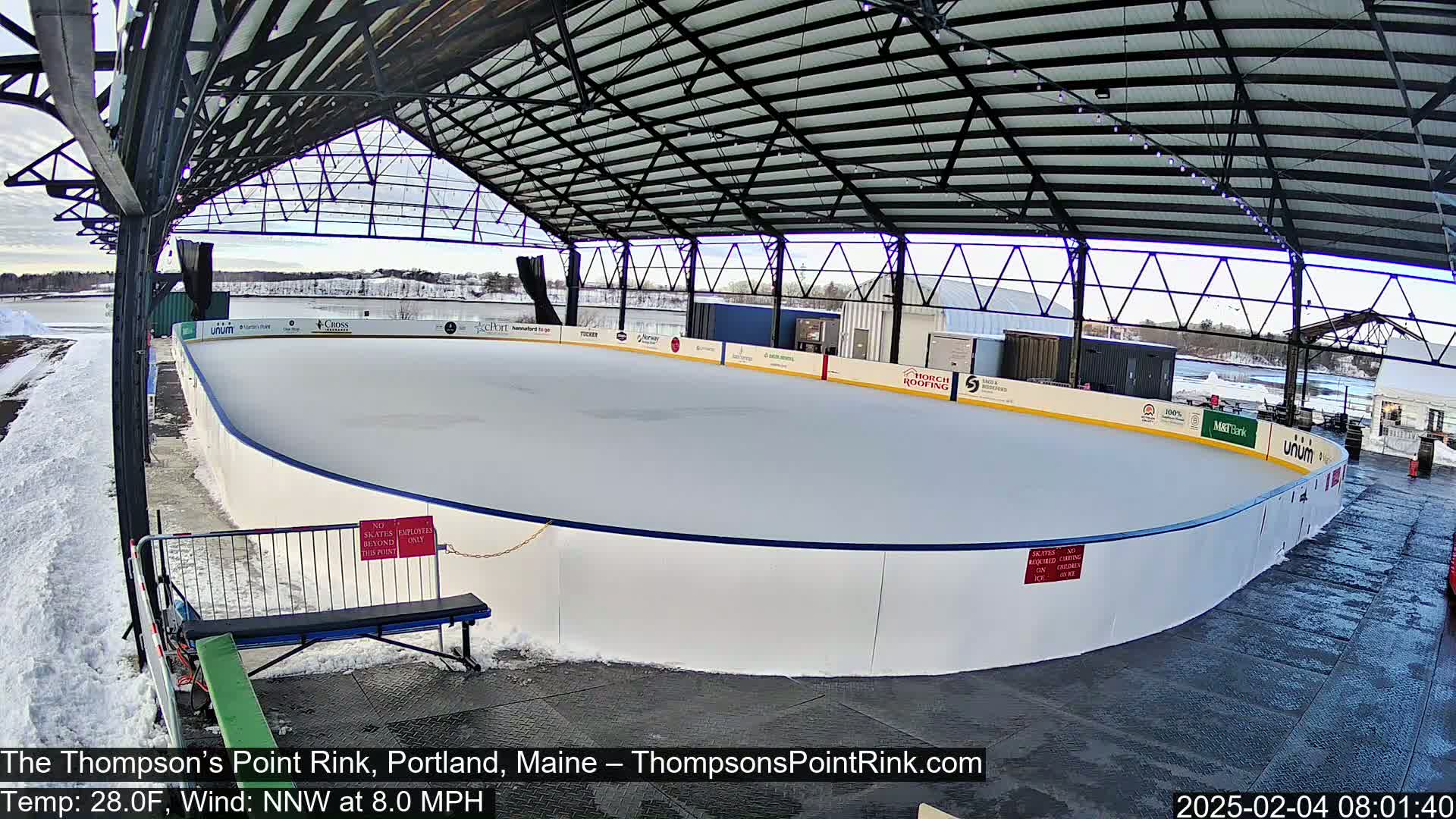 An outdoor ice rink under a metal roof, with snow on the ground and a partly cloudy sky.