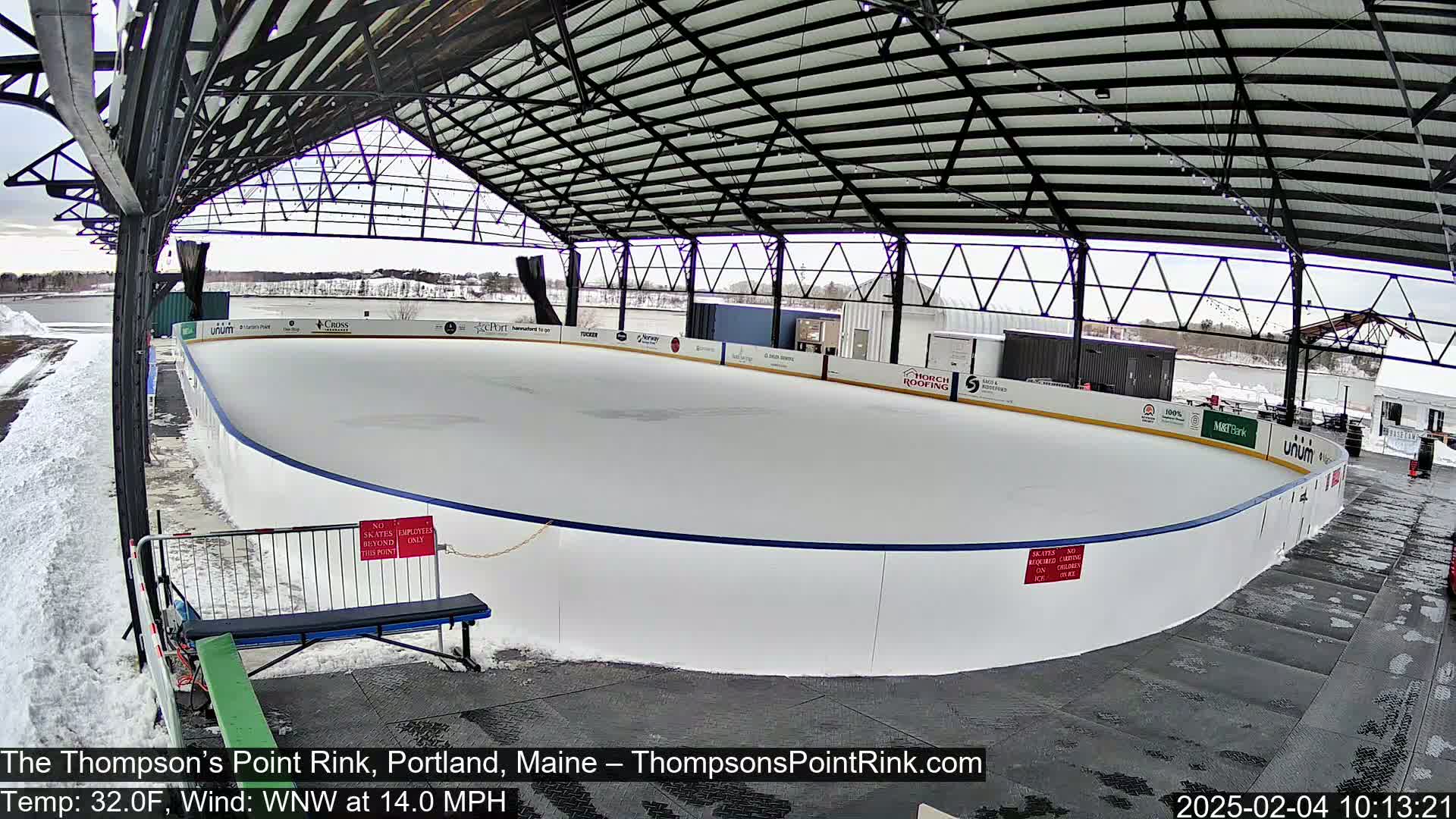 An outdoor ice rink, under a metal roof, is partially covered in snow with a 14 mph wind and a temperature of 32°F.