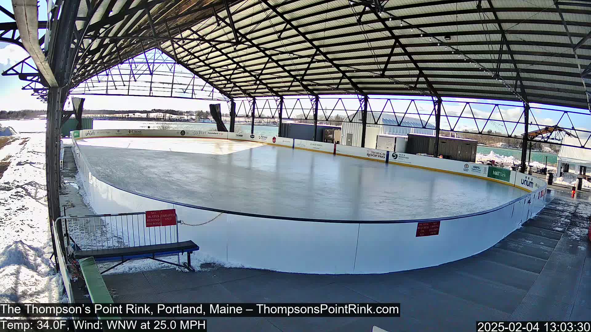 An outdoor ice rink, under a metal-framed roof, on a sunny day with a wind of 25 mph.