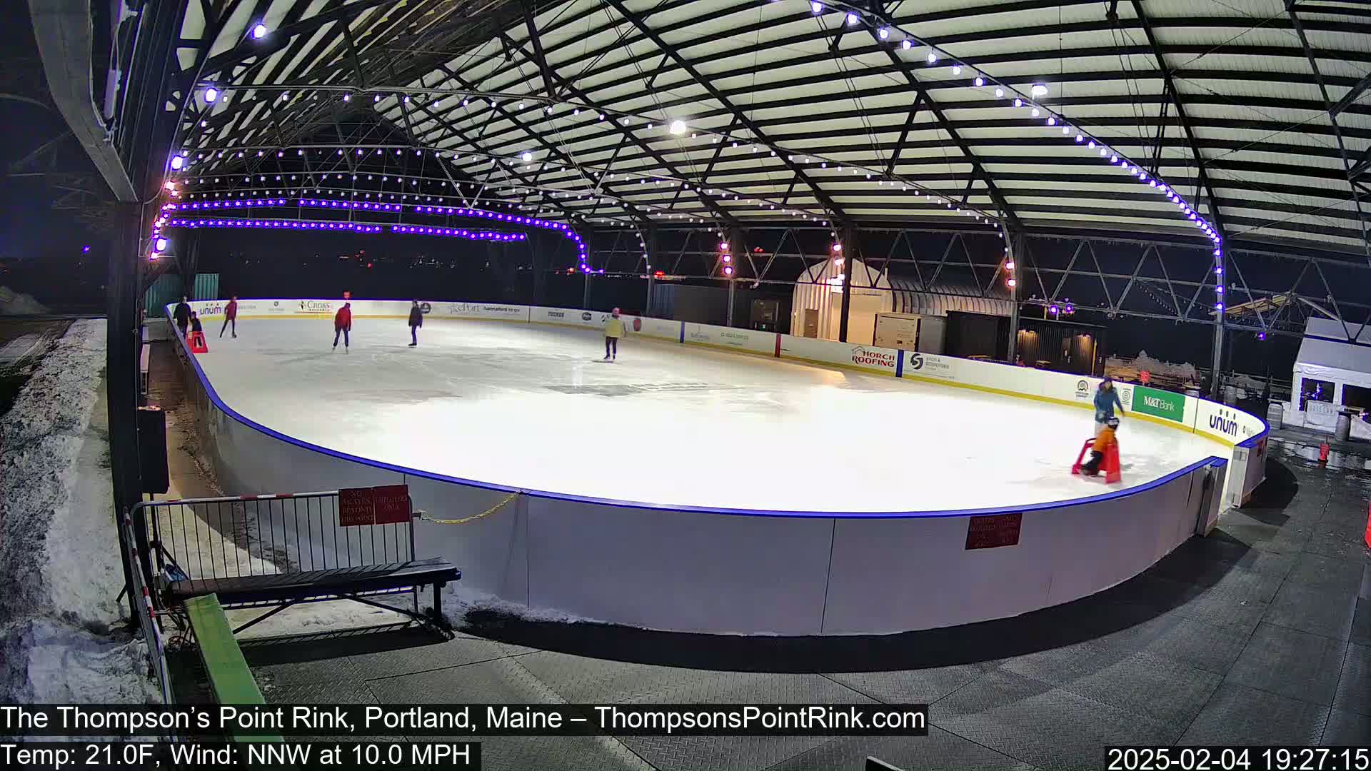 An outdoor ice rink at night, illuminated with purple lights, has several people ice skating, with some snow visible on the ground around the rink.