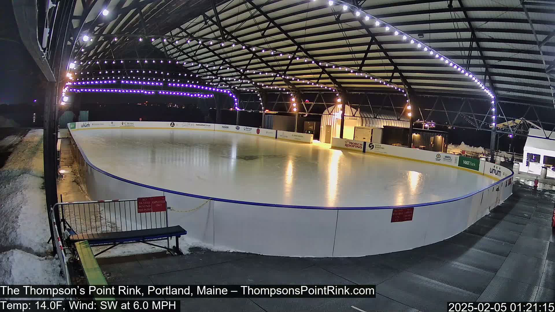 An outdoor ice rink, illuminated at night with string lights, is partially covered by a metal structure; there is snow on the ground.
