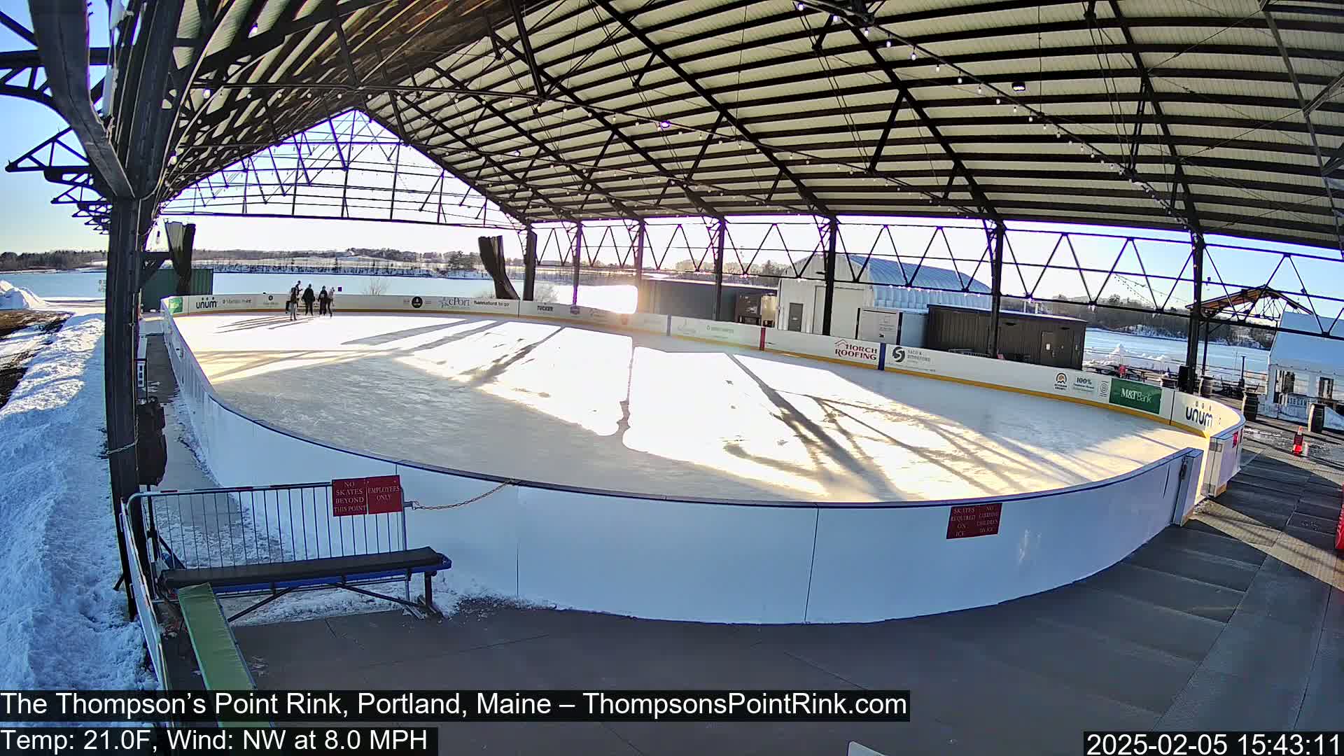 An outdoor ice rink, partially covered by a metal structure, is bathed in sunlight on a cold, windy day with snow on the ground.