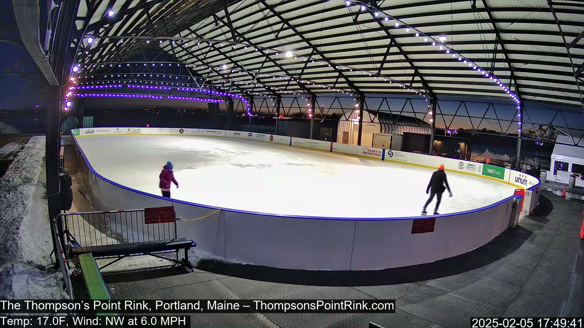 An outdoor ice rink, under a metal roof and illuminated by string lights, with two people ice skating at dusk in a cold, windy climate.