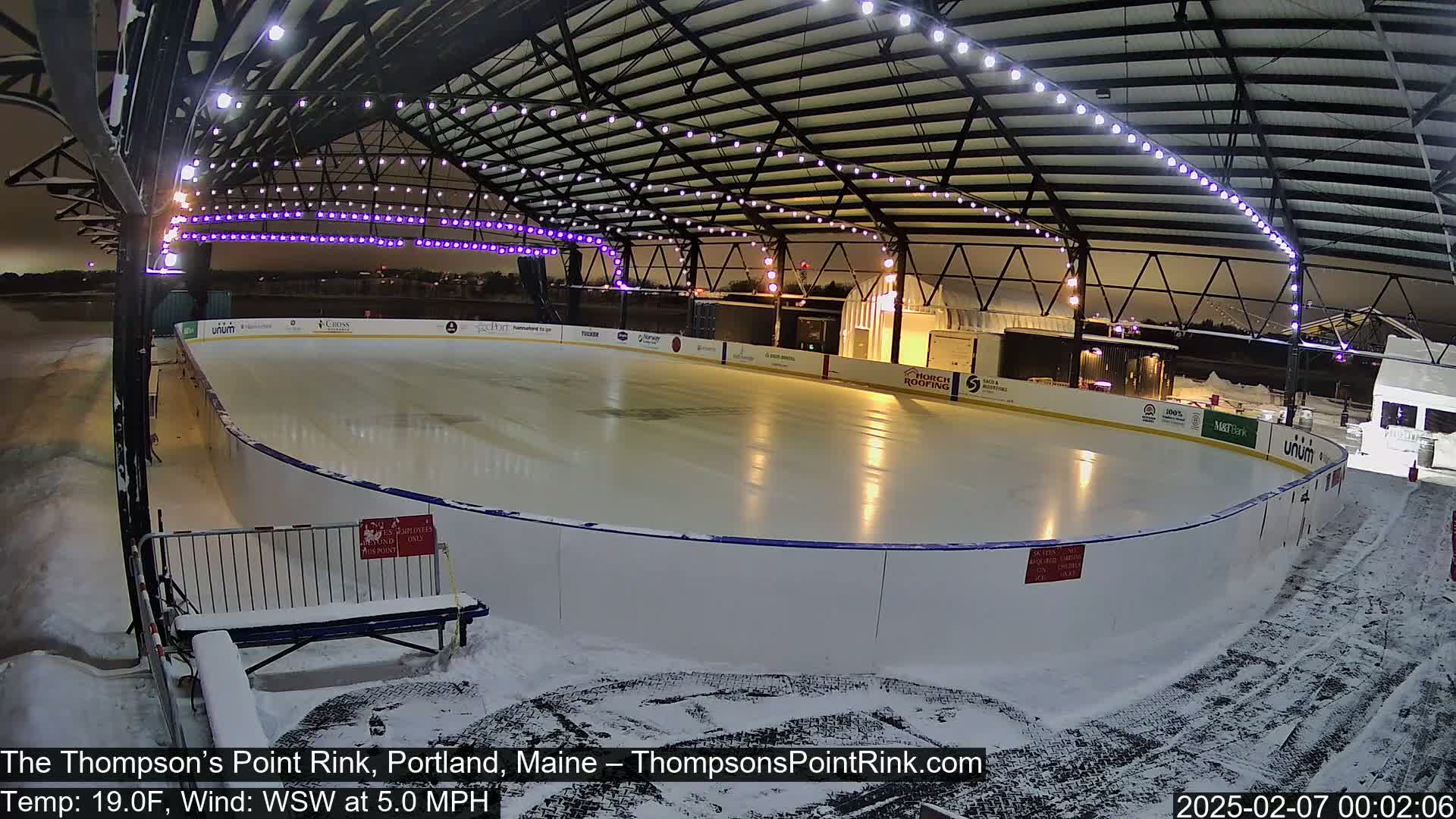 An outdoor ice rink, illuminated by overhead lights, is covered in a light layer of snow under a nighttime sky.