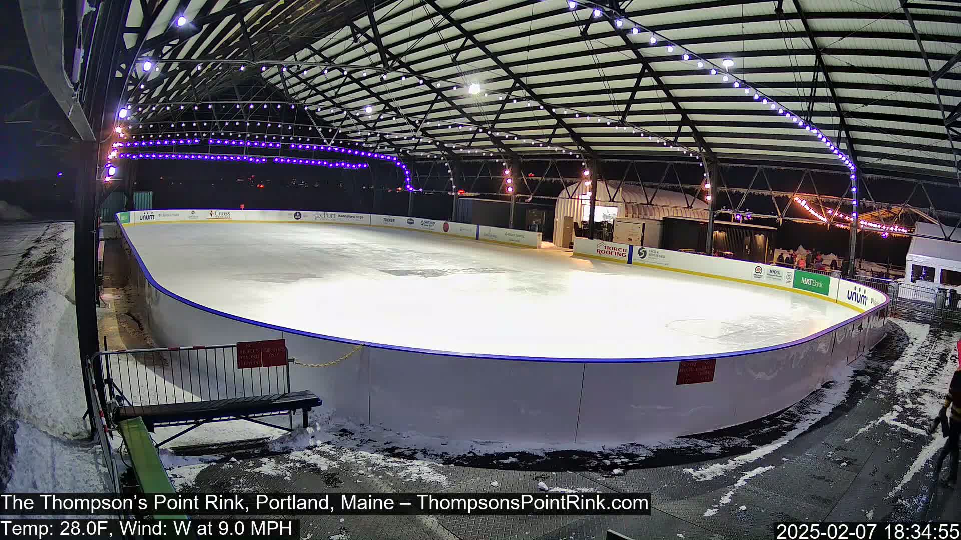 An outdoor ice rink is illuminated at night, surrounded by snow, under a metal roof structure with string lights.
