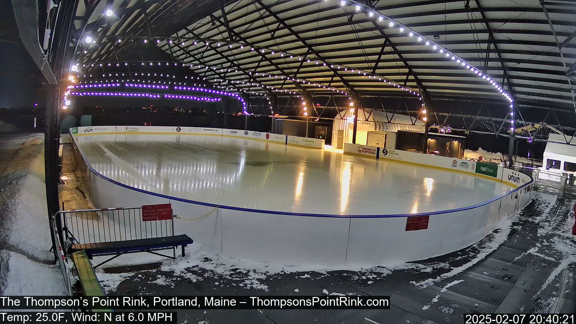 An outdoor ice rink at night, illuminated by lights, with some snow on the ground.