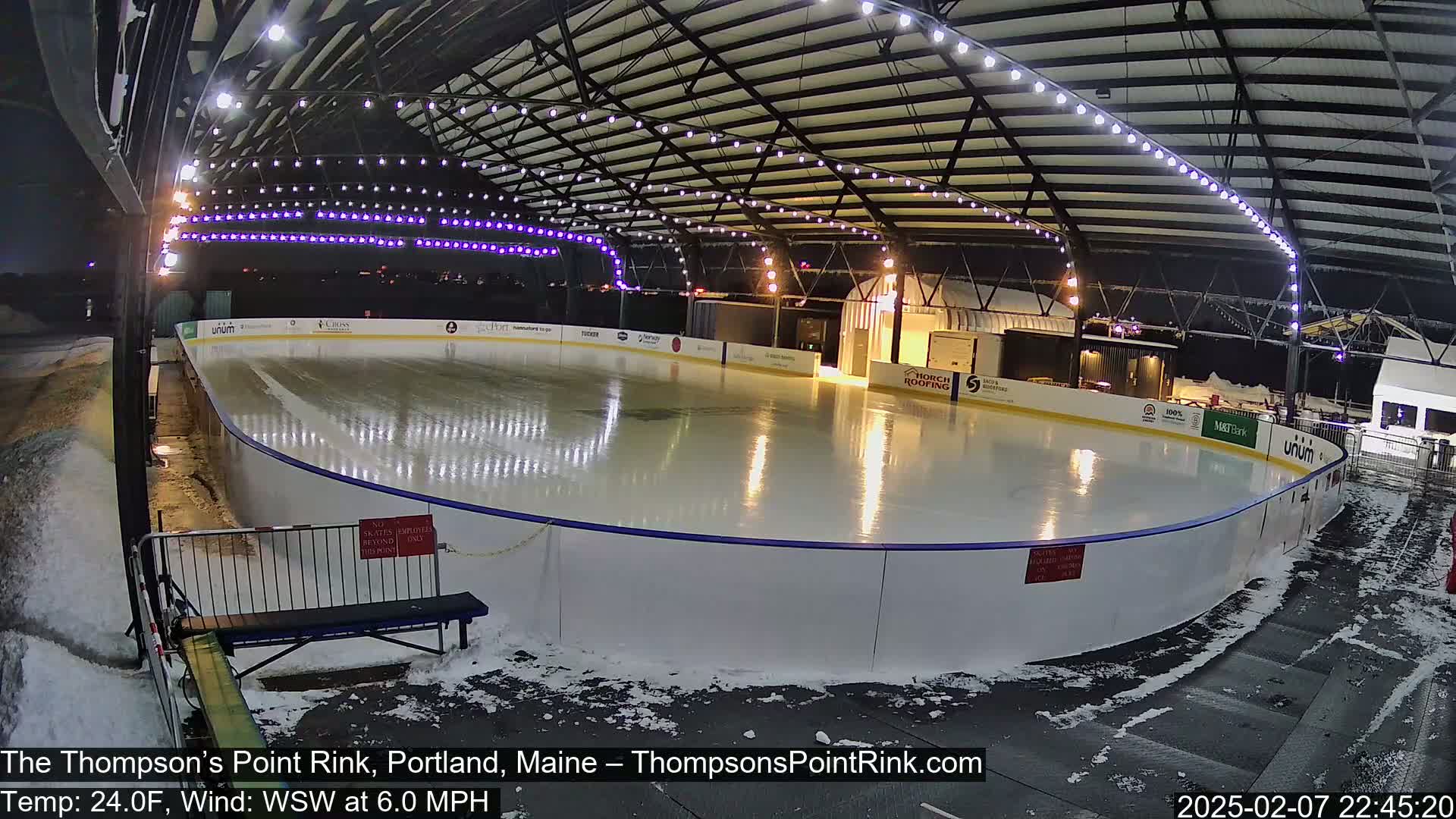 An outdoor ice rink, illuminated at night under a metal structure with string lights, is partially covered in snow.