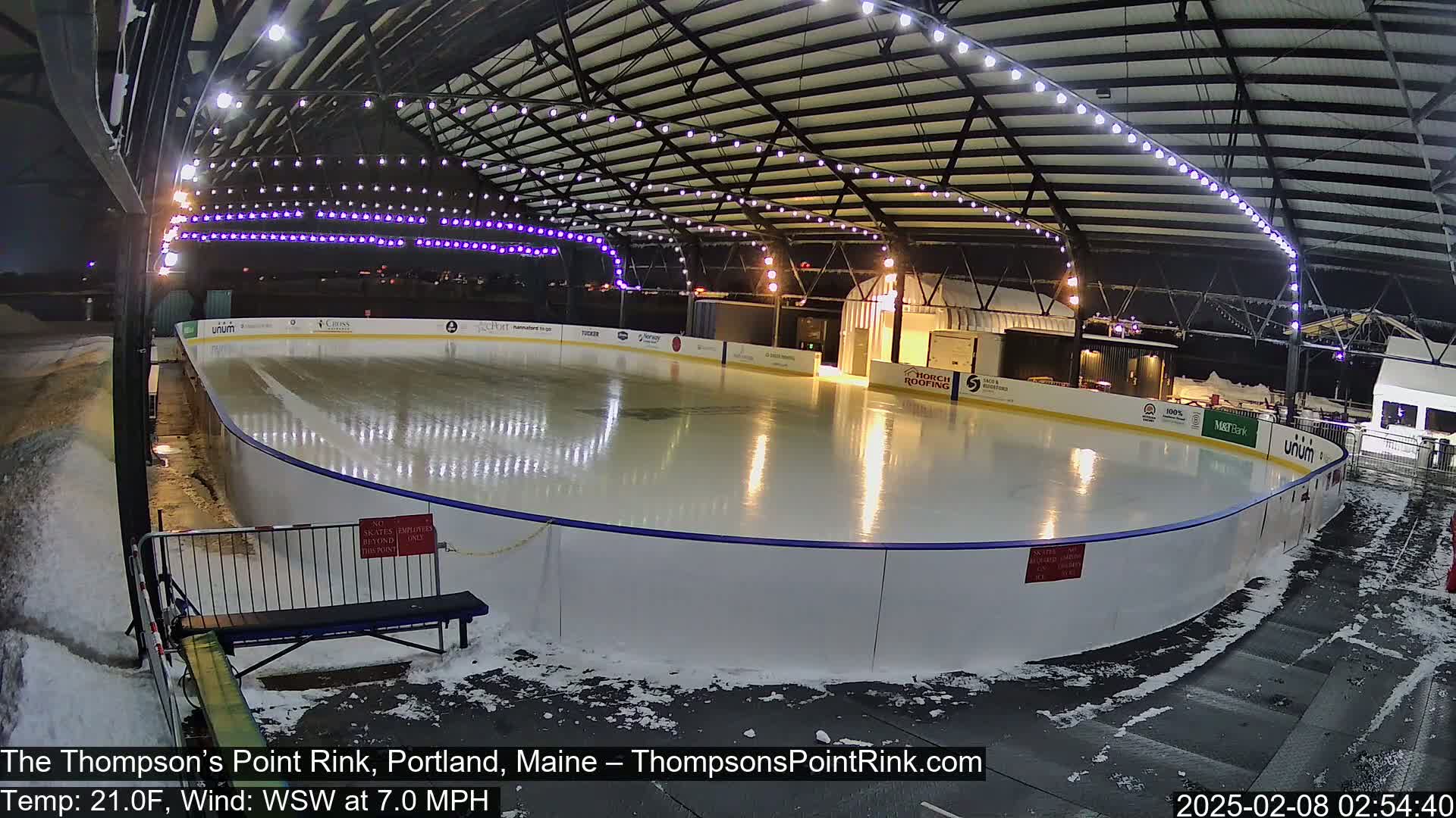 A brightly lit outdoor ice rink, partially covered in snow, is situated under a metal-framed structure with string lights at night.