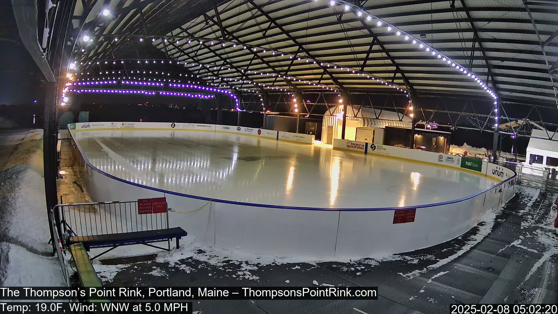 An outdoor ice rink, illuminated at night, is partially covered with snow, under a metal structure with string lights.