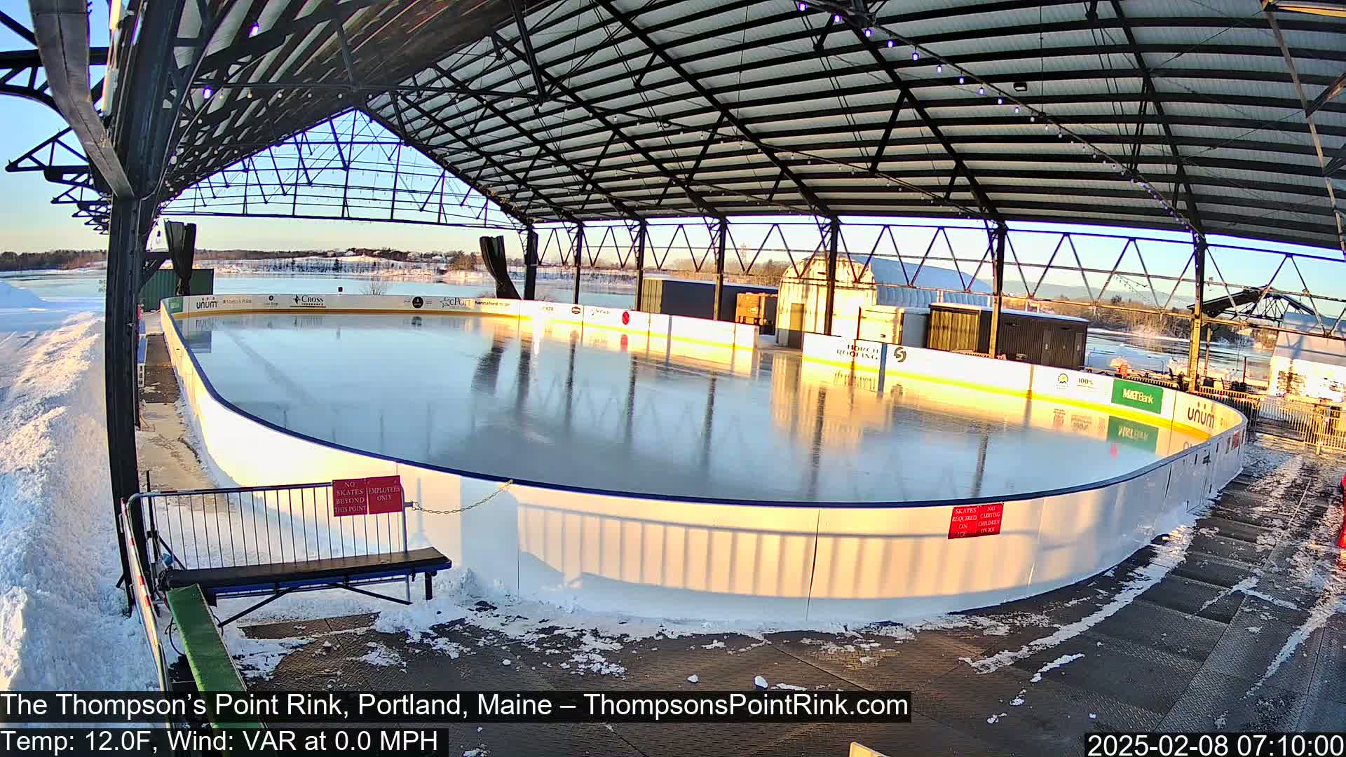 An outdoor ice rink sits under a metal structure on a sunny, cold day with snow on the ground.