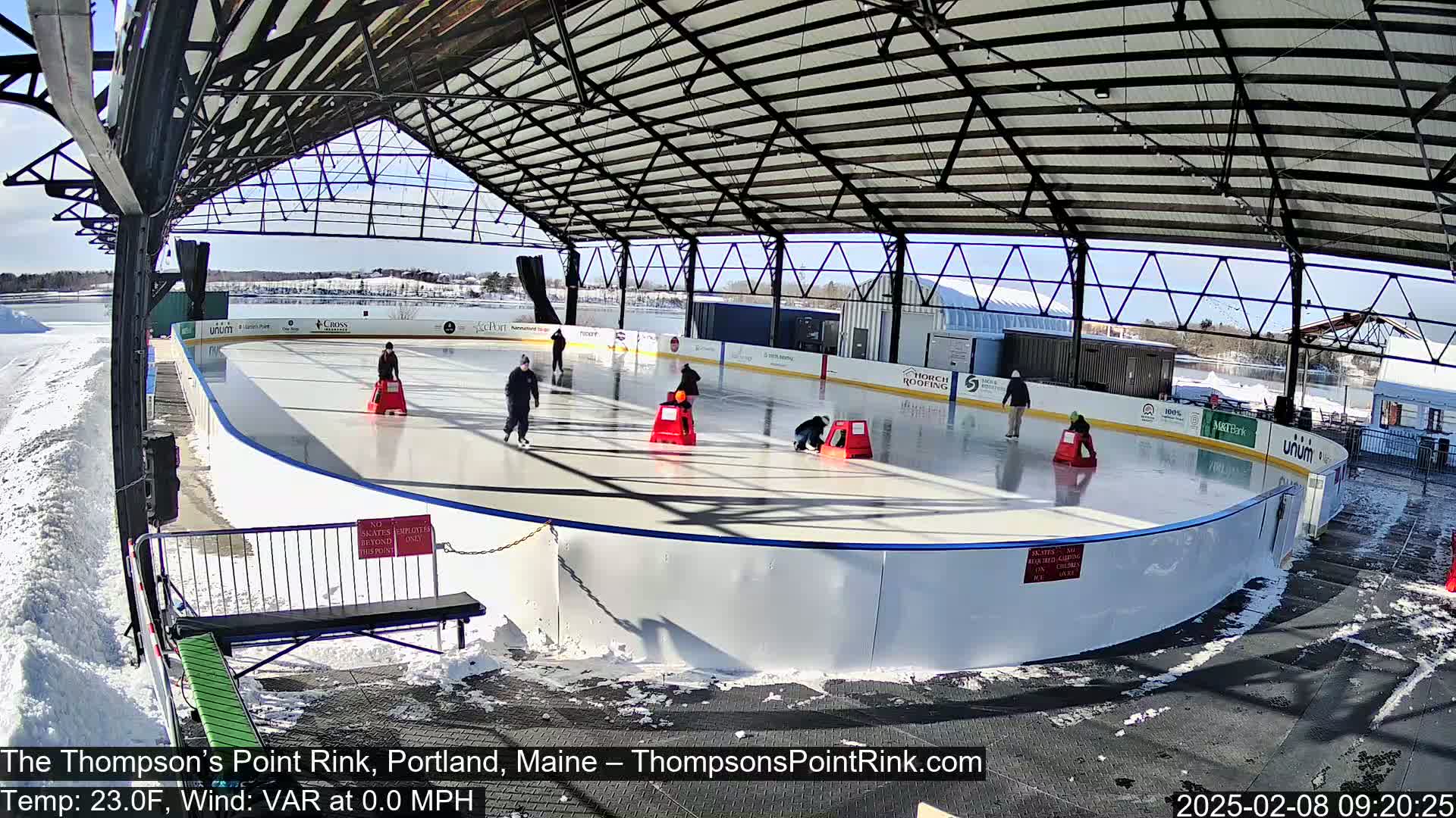 An outdoor ice rink, partially covered by a metal structure, is surrounded by snow on a sunny day.