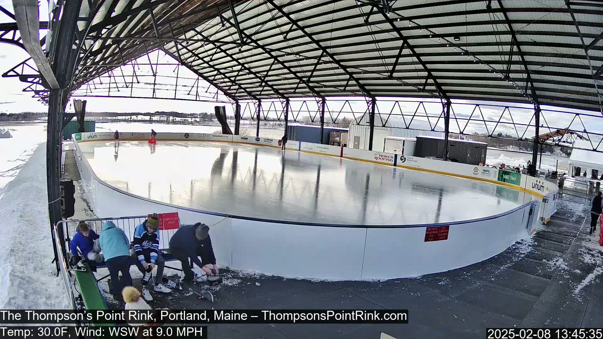 An outdoor ice rink under a metal structure on a partly sunny, cold day with a light wind, shows several people around the rink, some skating and others sitting near the side.