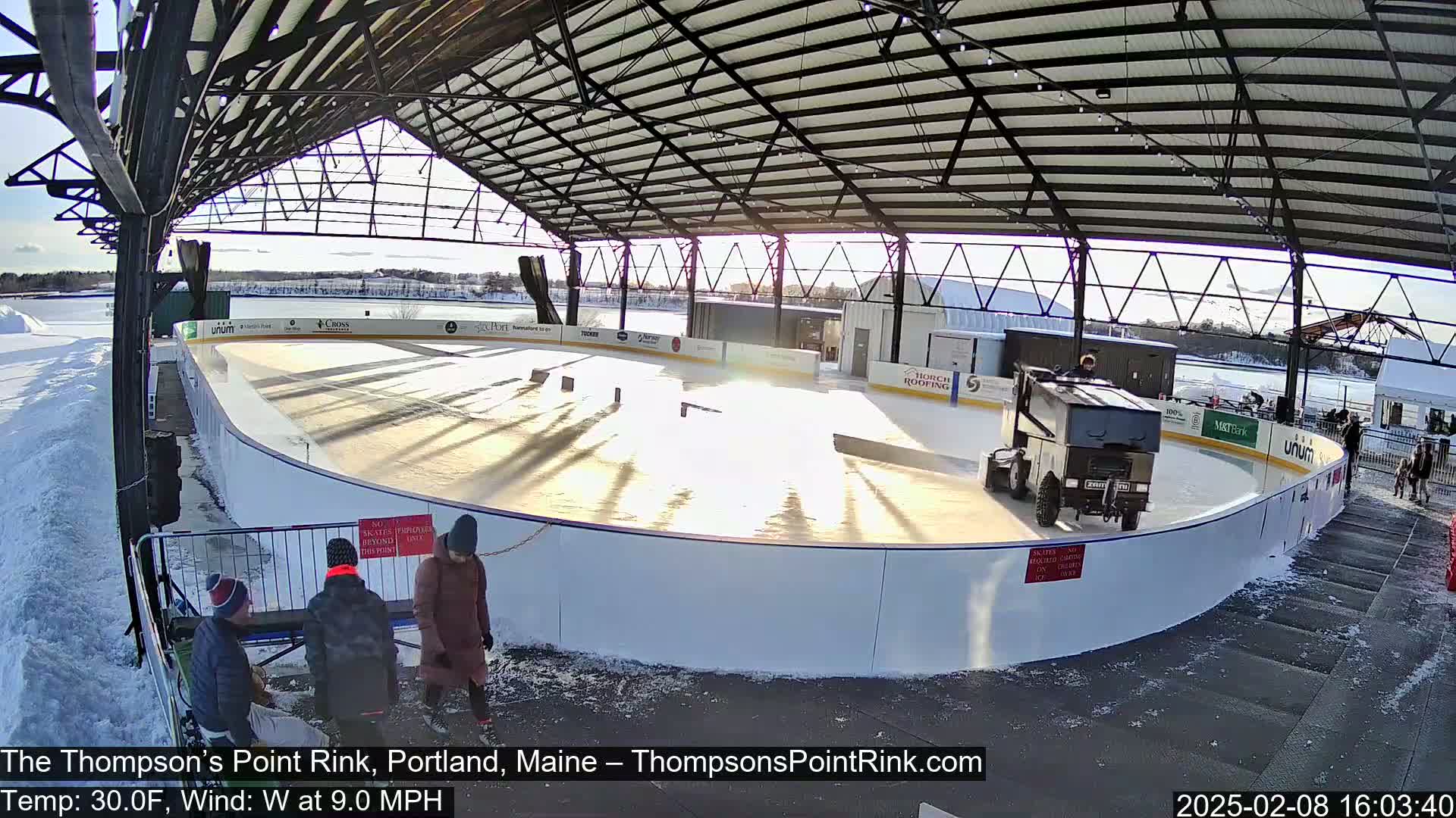 A sunny outdoor ice rink, partially covered by a metal structure, shows a Zamboni grooming the ice, with several people standing nearby on a snowy day.