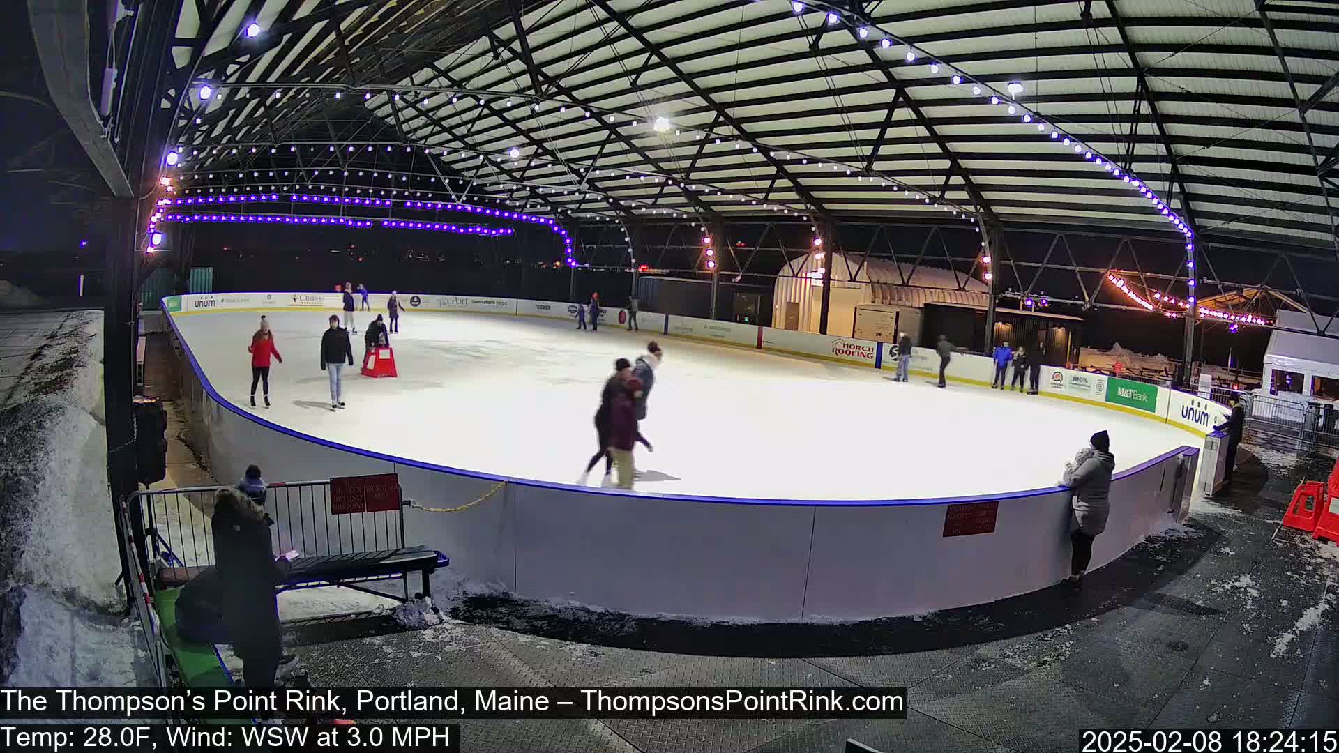 An outdoor ice rink at night, under a covered structure with string lights, is partially filled with ice skaters; there is snow on the ground.