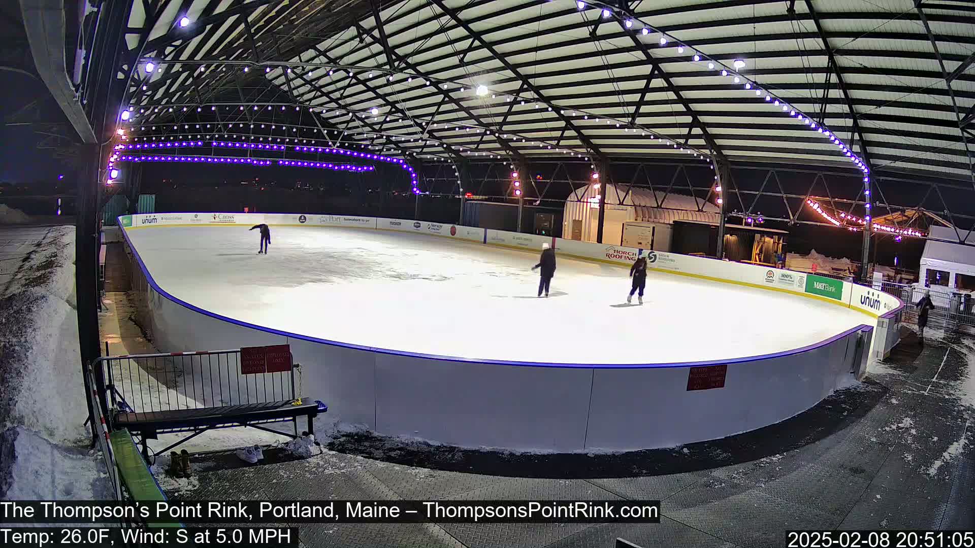 An outdoor ice rink at night, under a metal structure with lights, is partially covered in snow with several people ice skating.