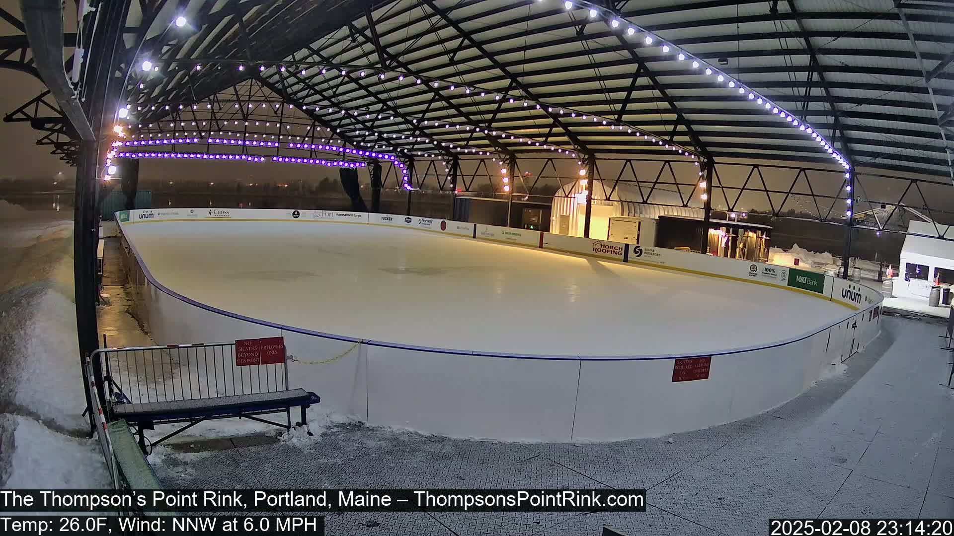 An outdoor ice rink under a covered structure is lightly snow-covered, with a temperature of 26 degrees Fahrenheit and a north-northwest wind at 6 miles per hour.