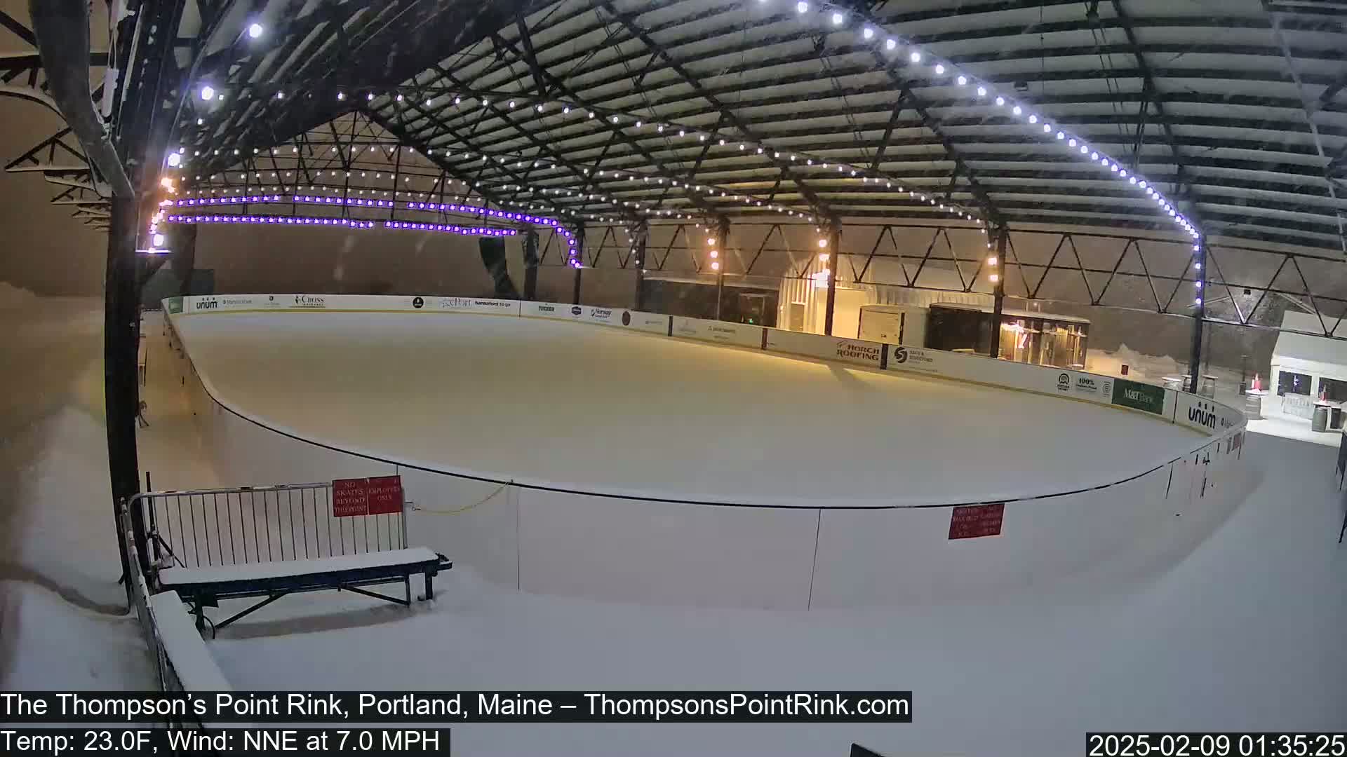 A snow-covered outdoor ice rink sits under a metal structure with string lights during a snowfall.