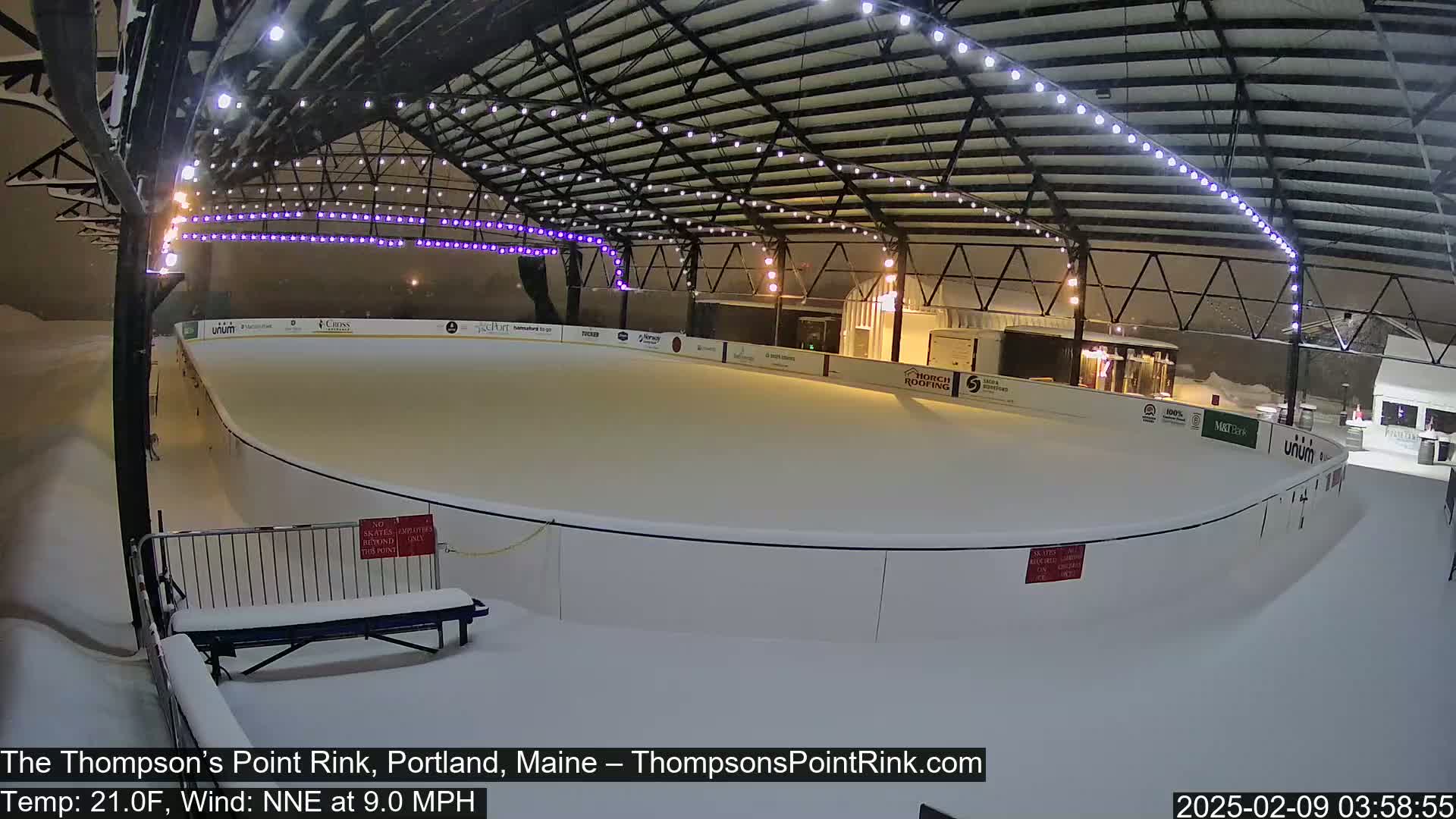 A snow-covered outdoor ice rink is illuminated under a metal structure with string lights during a snowfall, with a north-northeast wind.