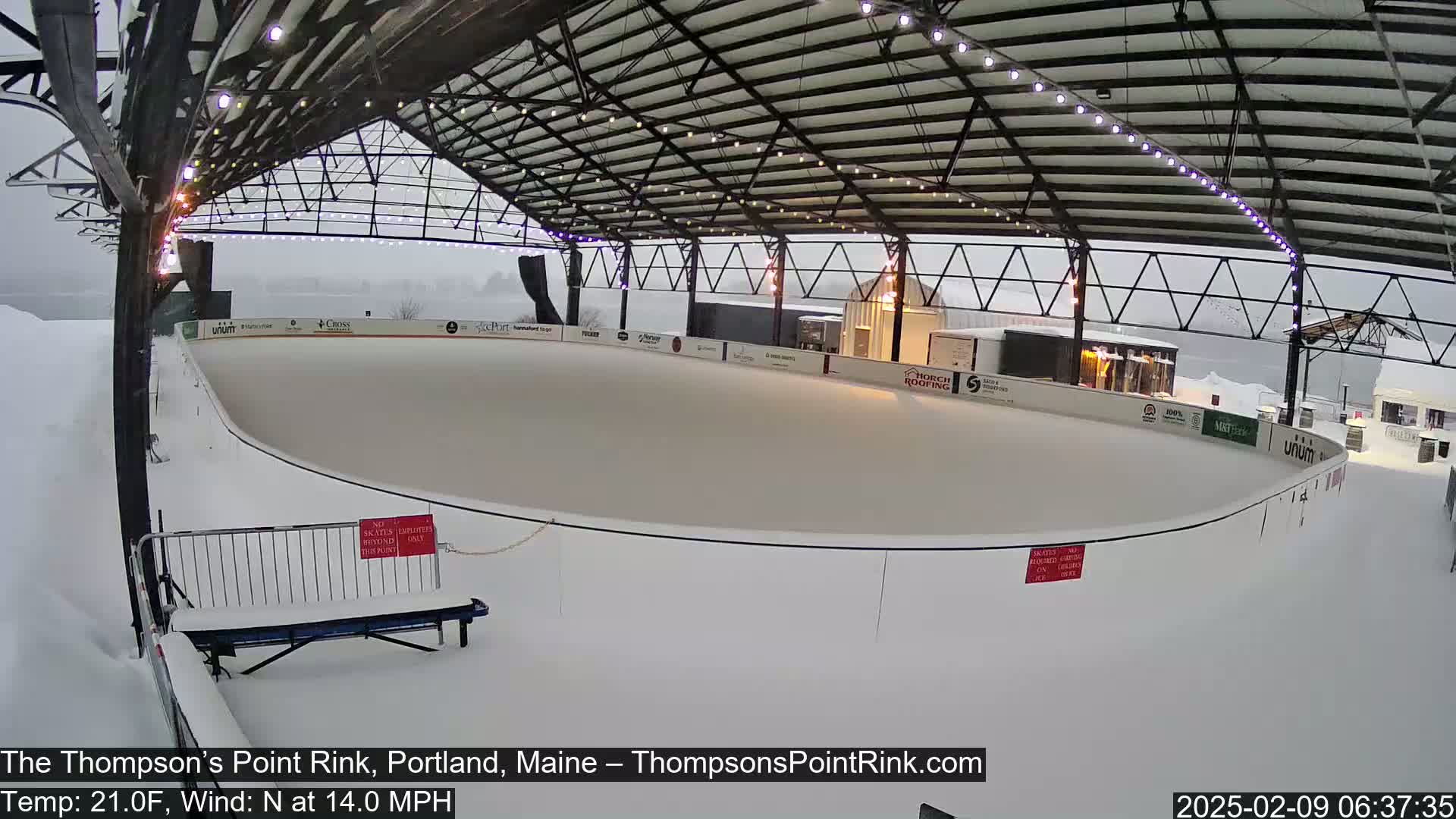 An outdoor ice rink covered by a metal structure is lightly snowing.