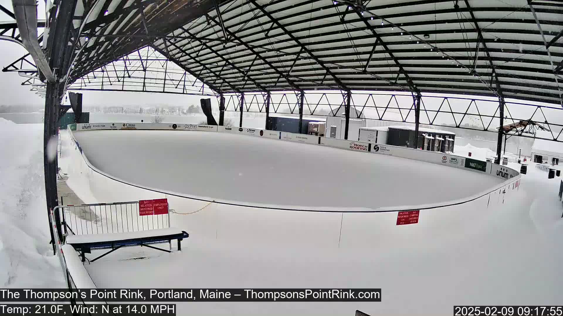 A snow-covered outdoor ice rink sits under a metal-framed shelter during a snowfall.