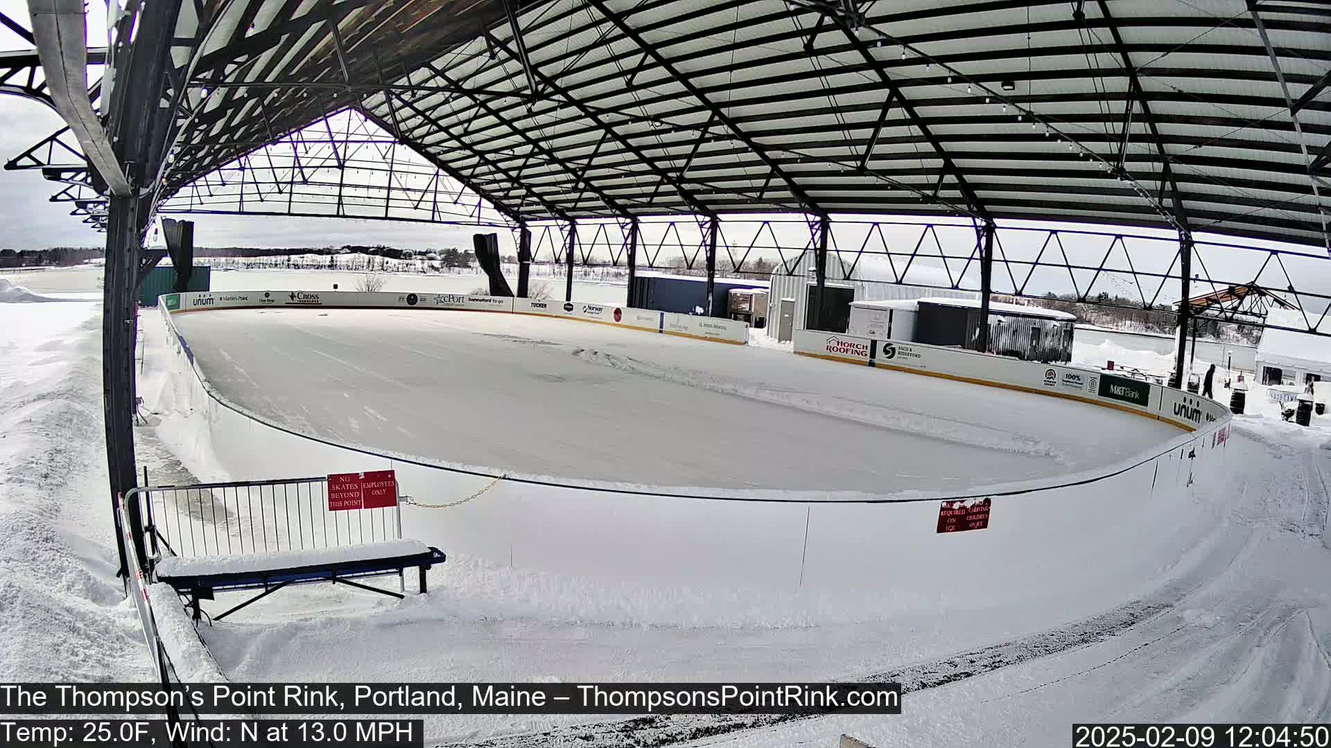 An outdoor ice rink covered by a metal structure is surrounded by snow, under a cloudy sky.