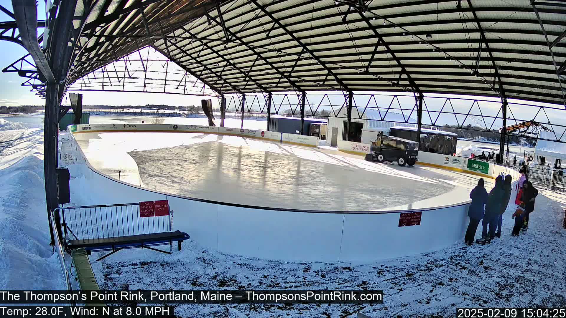 An outdoor ice rink, covered by a metal structure, is partially surrounded by snow on a sunny day with a north wind.