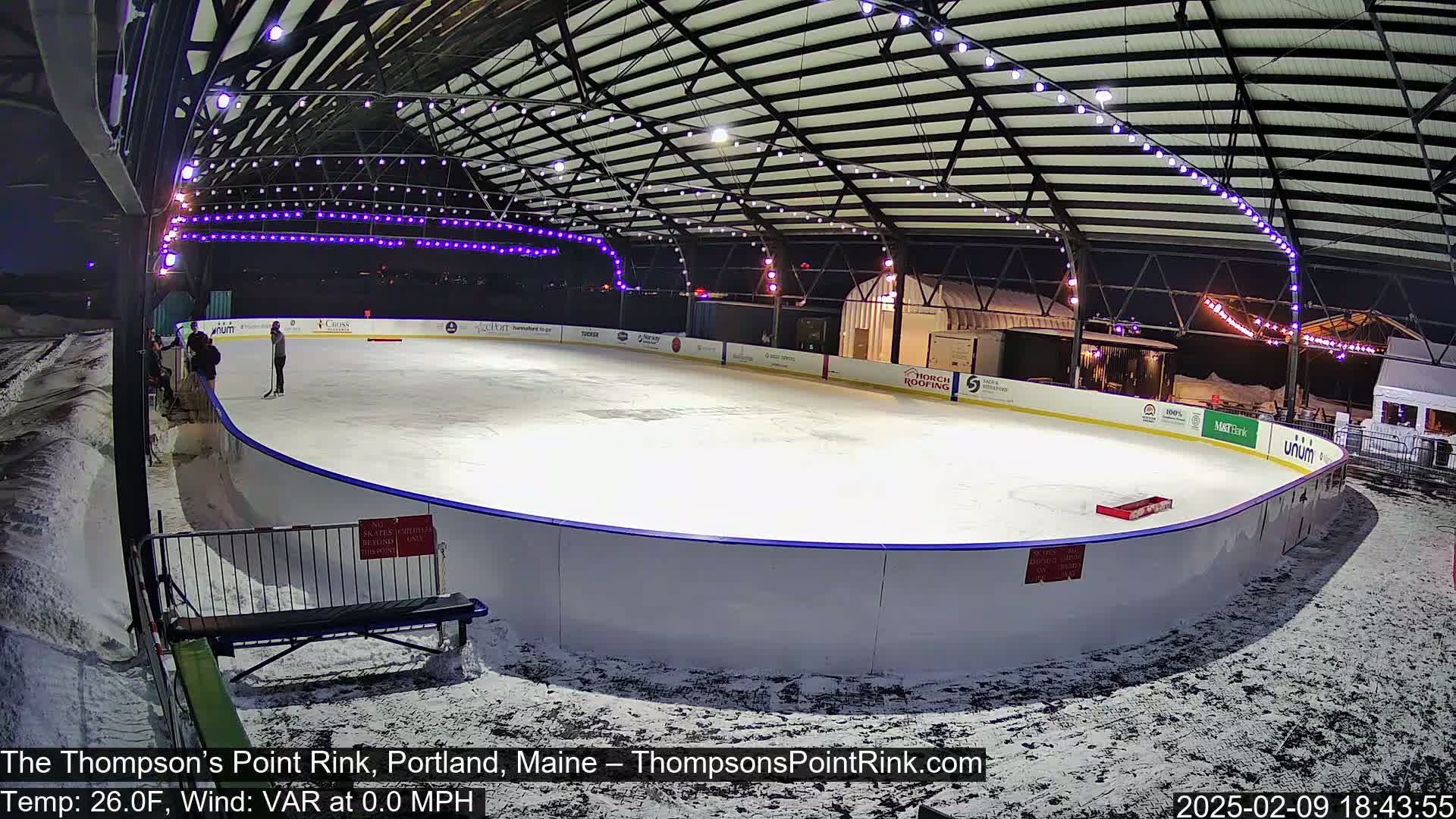 An outdoor ice rink, lit at night under a metal structure, is mostly empty with a few people skating and some snow on the ground.