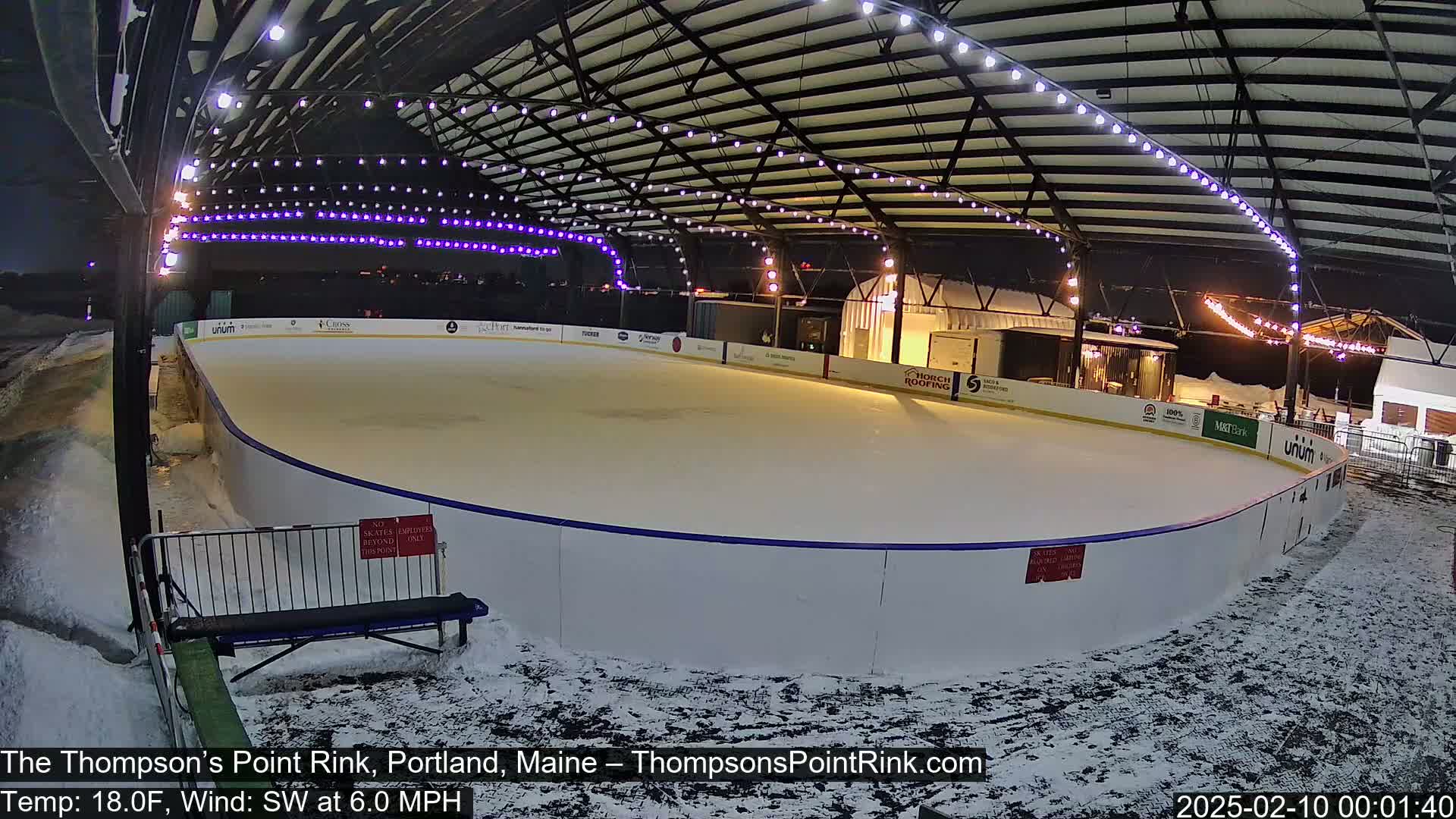 An outdoor ice rink, lit with string lights, is partially covered by a metal structure; snow is on the ground around the rink.