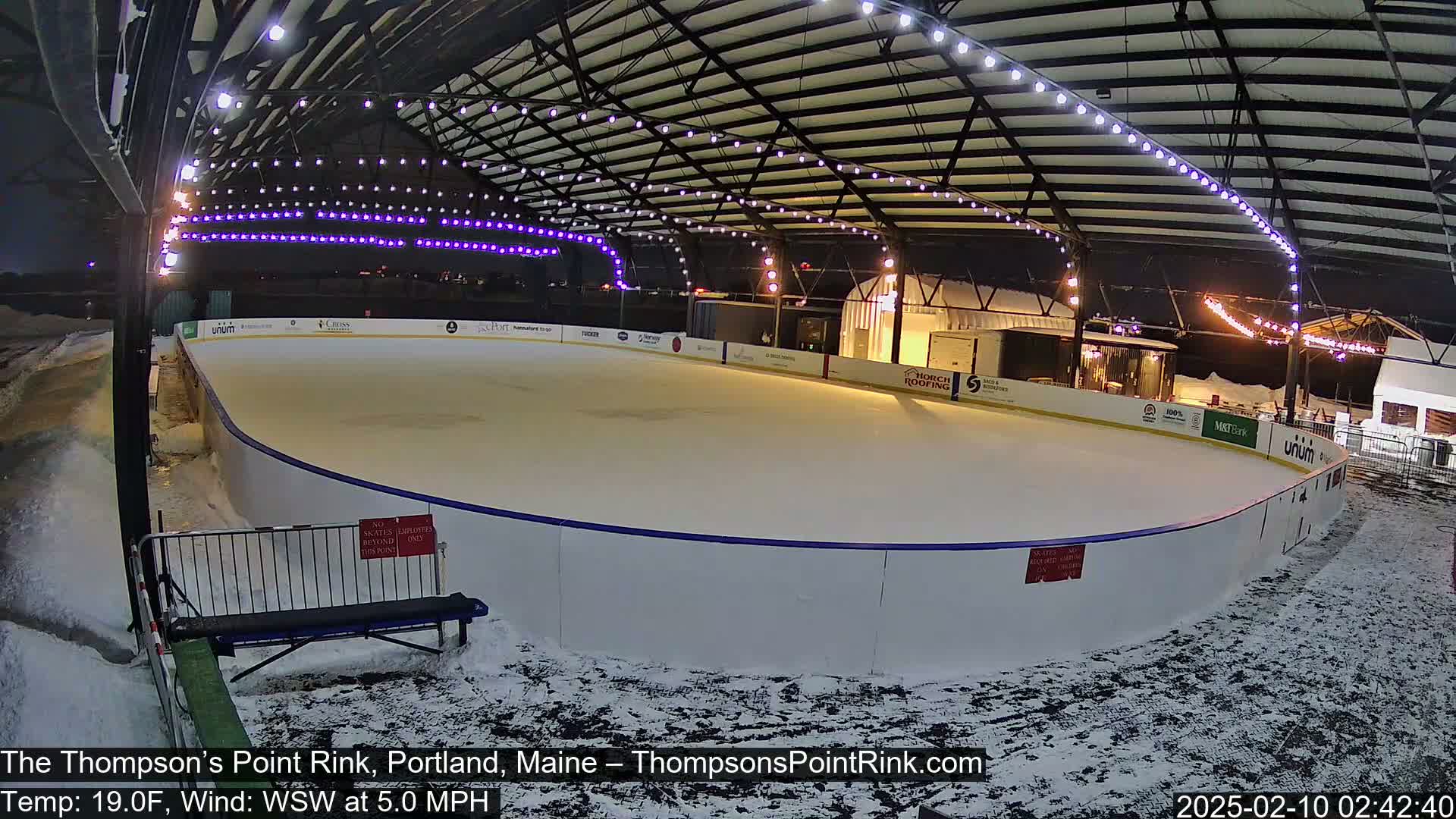 An outdoor ice rink, illuminated at night under a metal structure, is surrounded by snow.