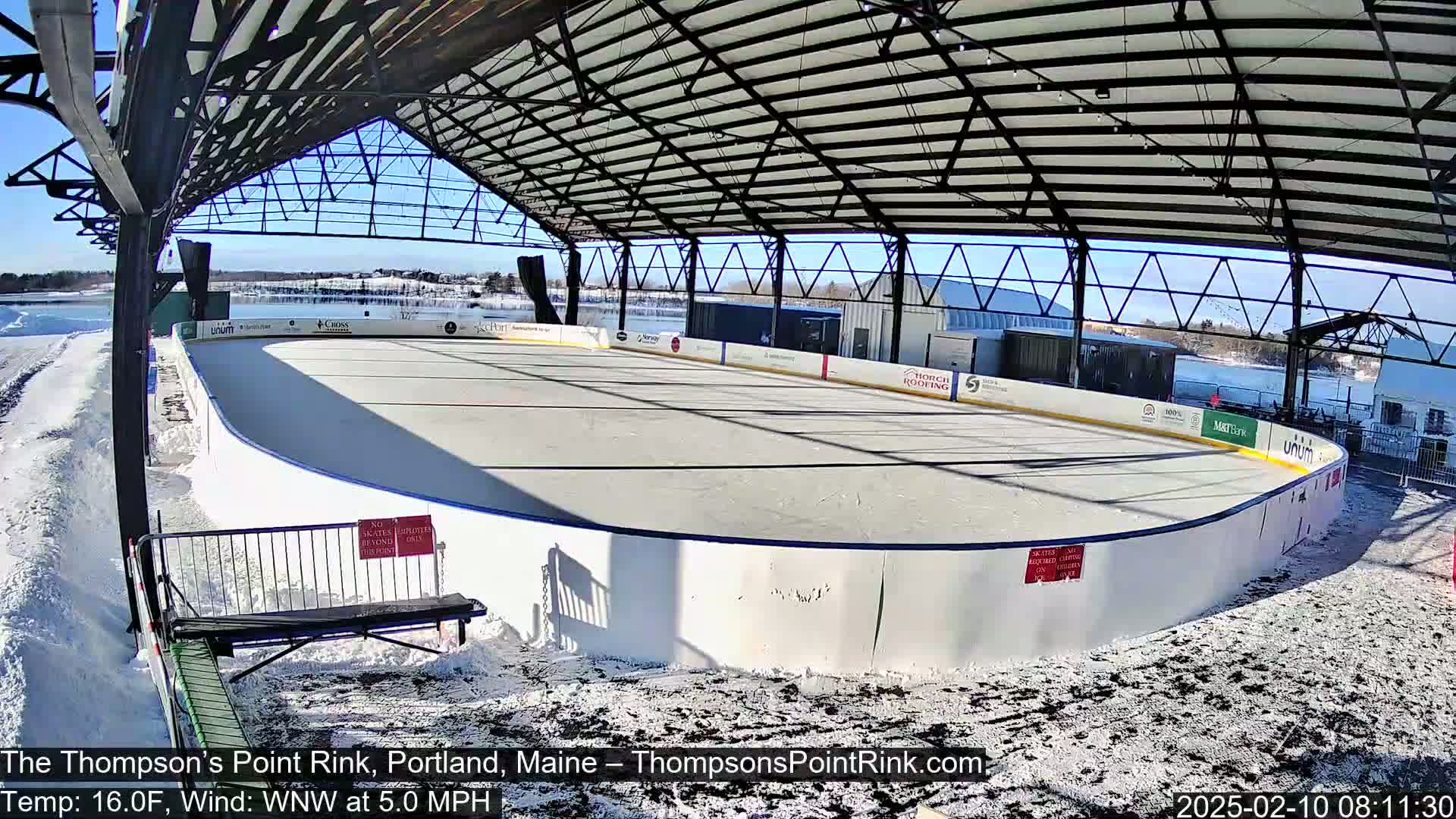 An outdoor ice rink, covered by a metal structure, is surrounded by snow on a sunny day with a light wind.