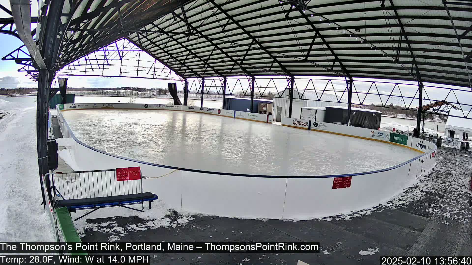 An outdoor ice rink, covered by a metal structure, is partially surrounded by snow in windy, 28-degree Fahrenheit weather.
