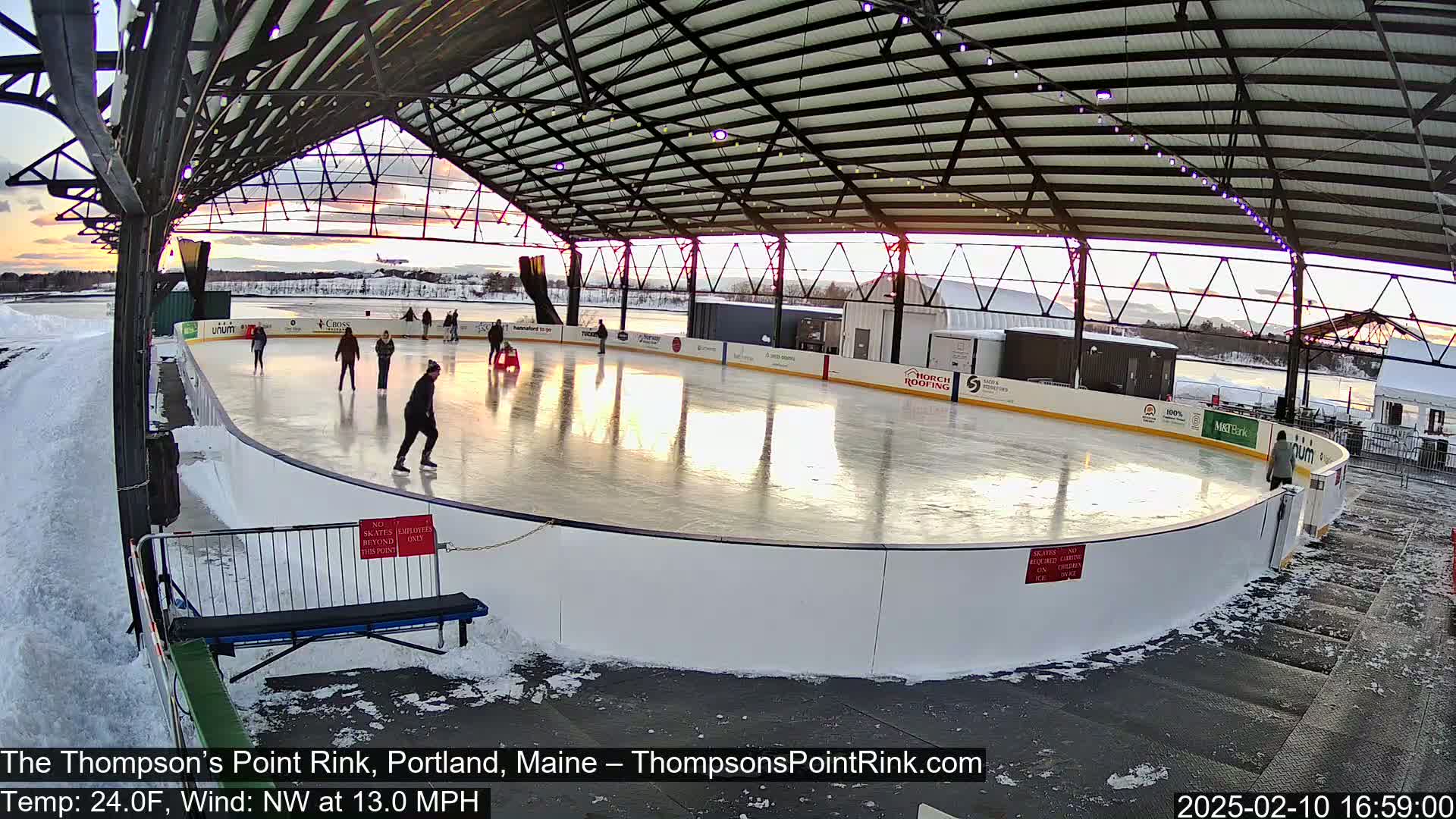 An outdoor ice rink, under a metal framework, is partially surrounded by snow, with several people ice skating as the sun sets.
