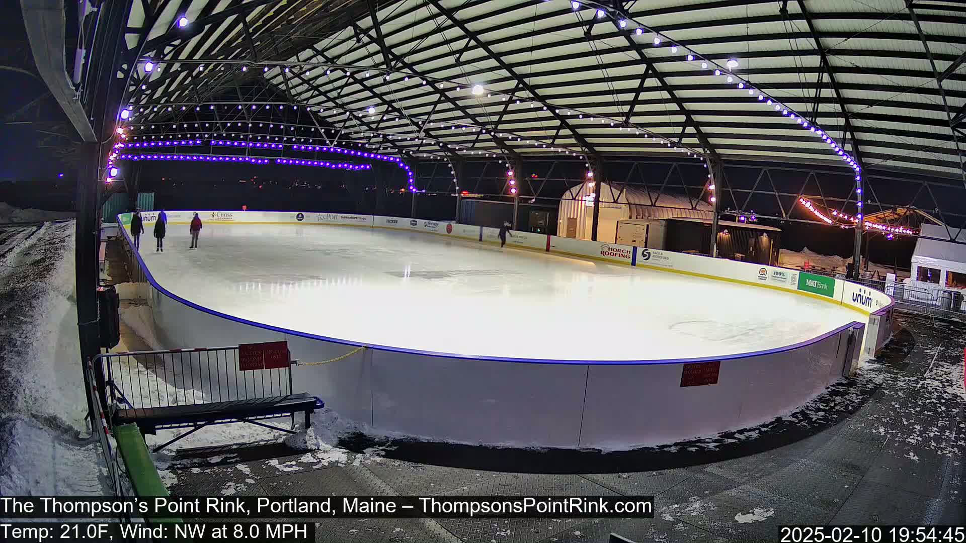 An outdoor ice rink, lit at night under a metal roof structure, with a few people ice skating and snow on the ground around the rink.