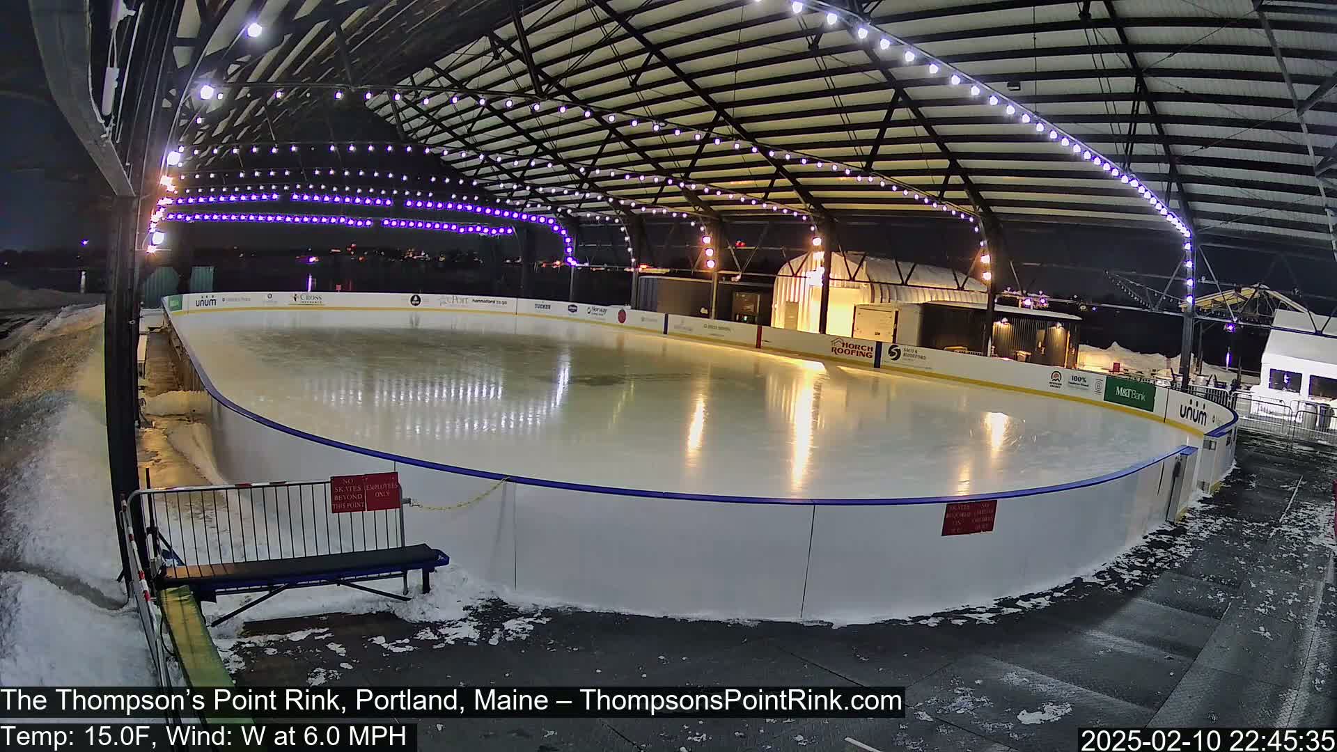 An outdoor ice rink is illuminated at night, surrounded by snow and under a covered structure with string lights.
